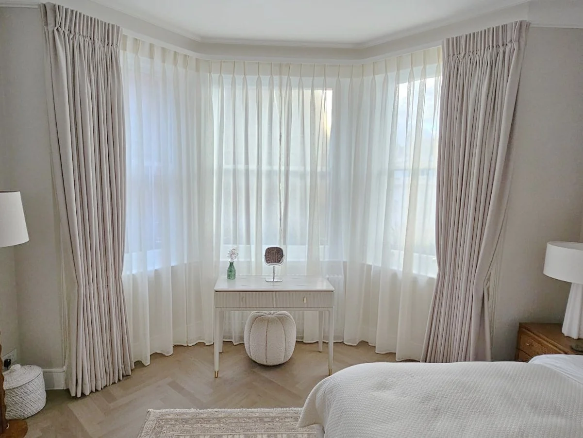 Bright bedroom corner with large bay window dressed in sheer white curtains and light beige drapes, a white writing desk with a small decorative mirror, a stool, and lamps on bedside tables.