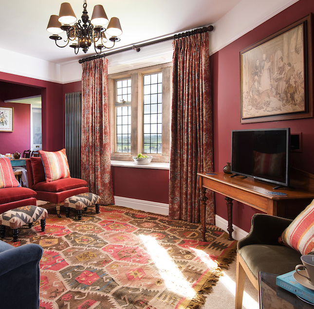 A cozy living room with red walls, floral curtains, patterned area rug, vintage chairs, a wooden TV stand, and a small window letting in sunlight.