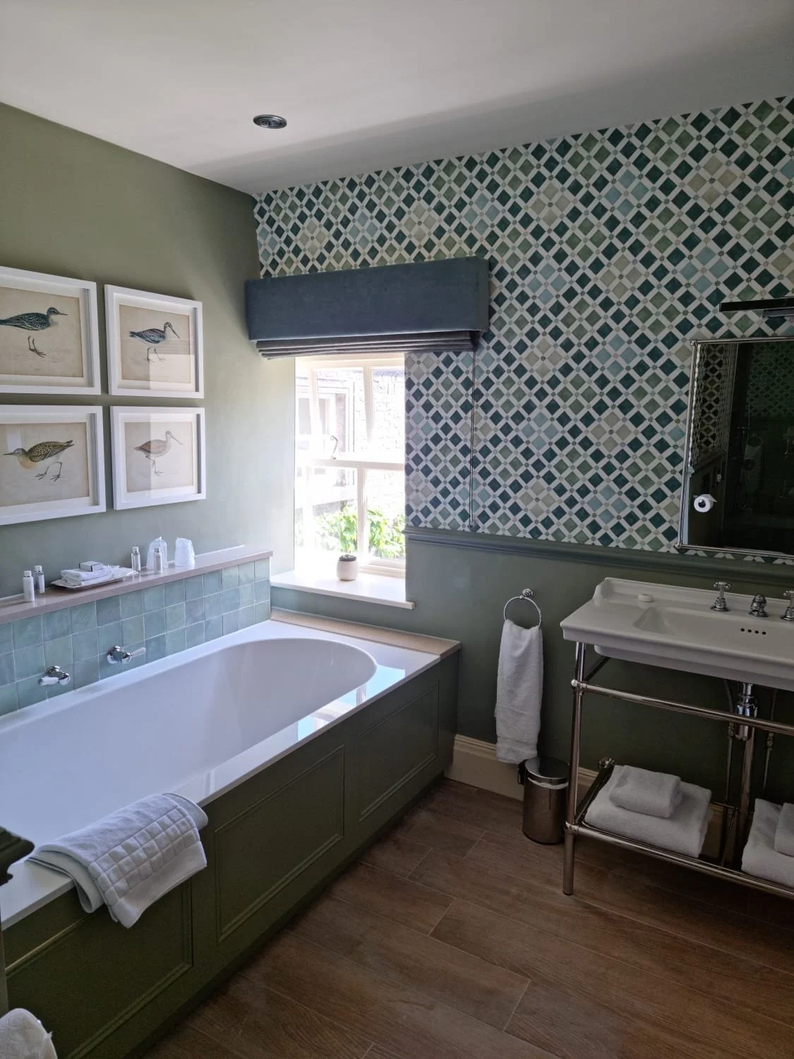 Bathroom with green walls, a built-in bathtub, framed bird artwork, a window with a blue valance, mosaic tile accent wall, sink with a towel rack, and wooden flooring.