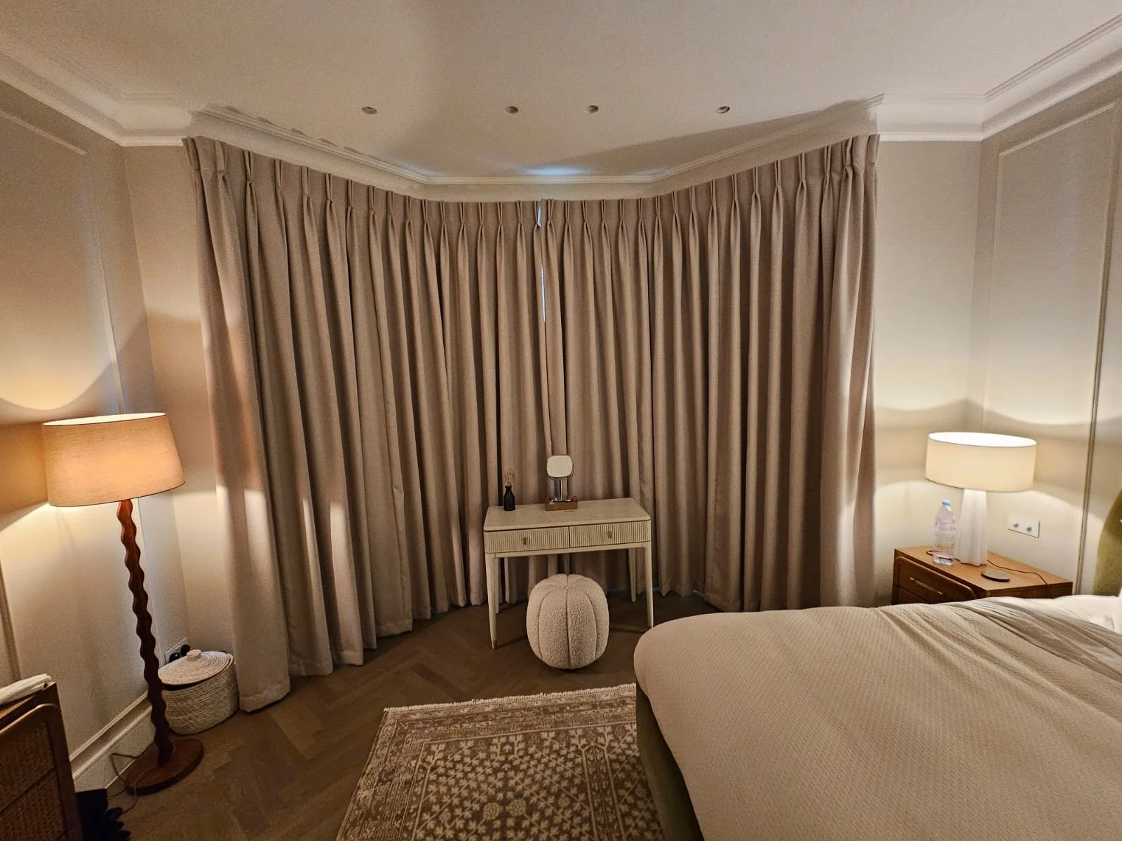 A hotel room with beige curtains, a bed, two lamps on side tables, a small bedside table with a mirror, and a decorative ottoman.