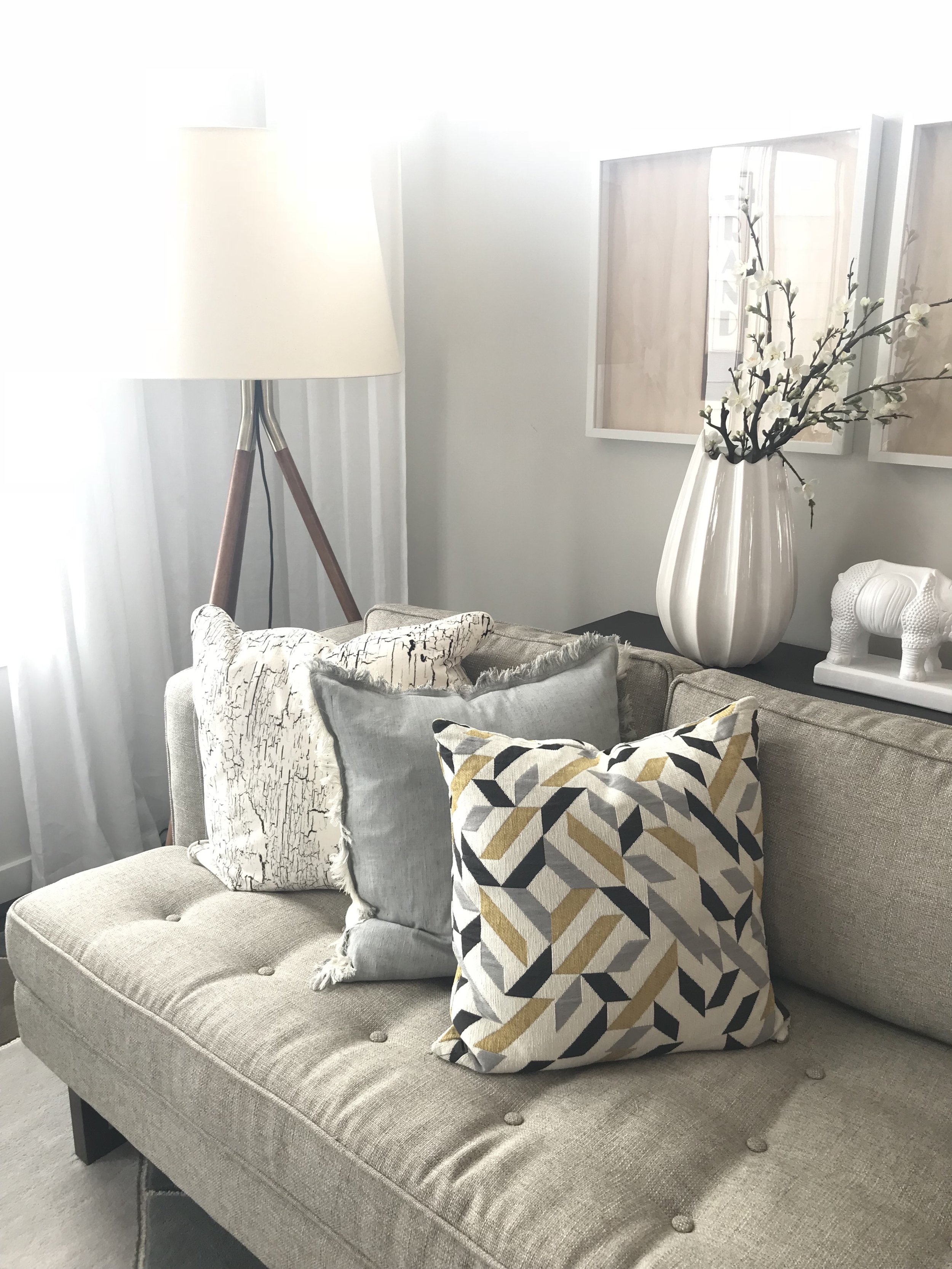 Living room corner with beige sofa, decorative pillows, white vase with branches, elephant figurine, tall lamp, light curtains, wall art