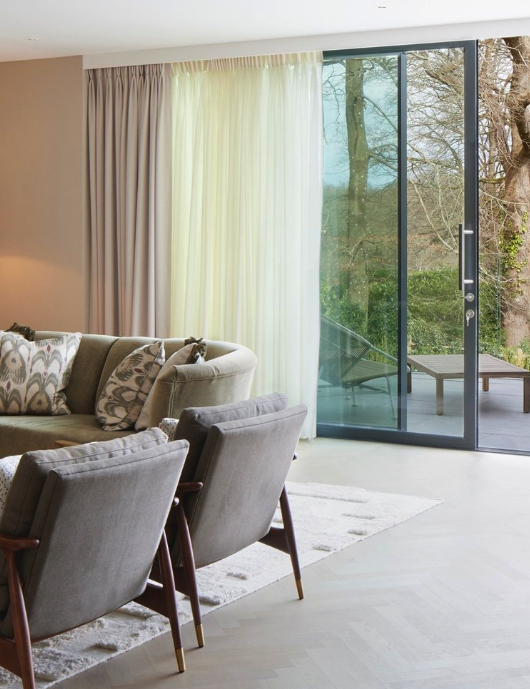 Living room with beige sofa, gray armchairs, curtains, and a sliding glass door leading to an outdoor patio with trees