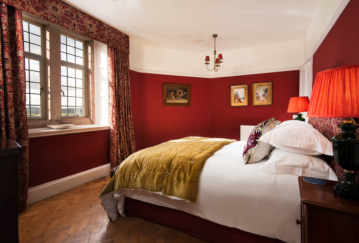 A bedroom with a large window, red walls, a bed with white sheets, a green bedspread, and decorative pillows. Red lamps are on bedside tables, and three paintings hang on the wall.
