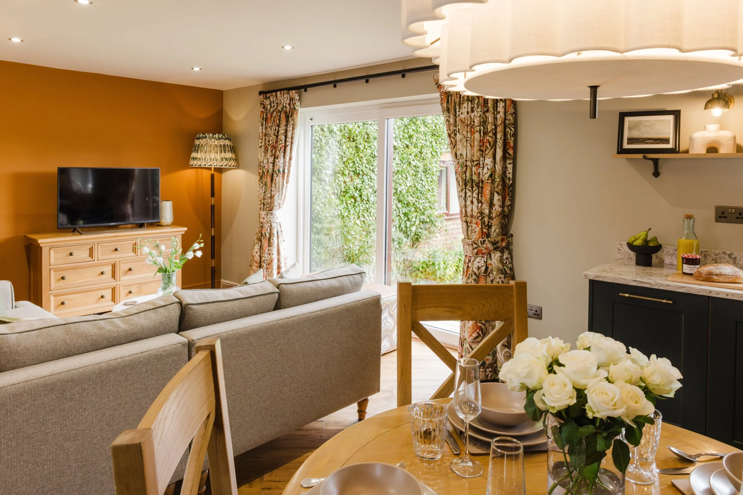 Living and dining room with a beige sofa, wooden sideboard with a TV, table with white roses, glassware, and tableware, window with floral curtains, and a kitchen area with dark cabinets and a marble countertop.