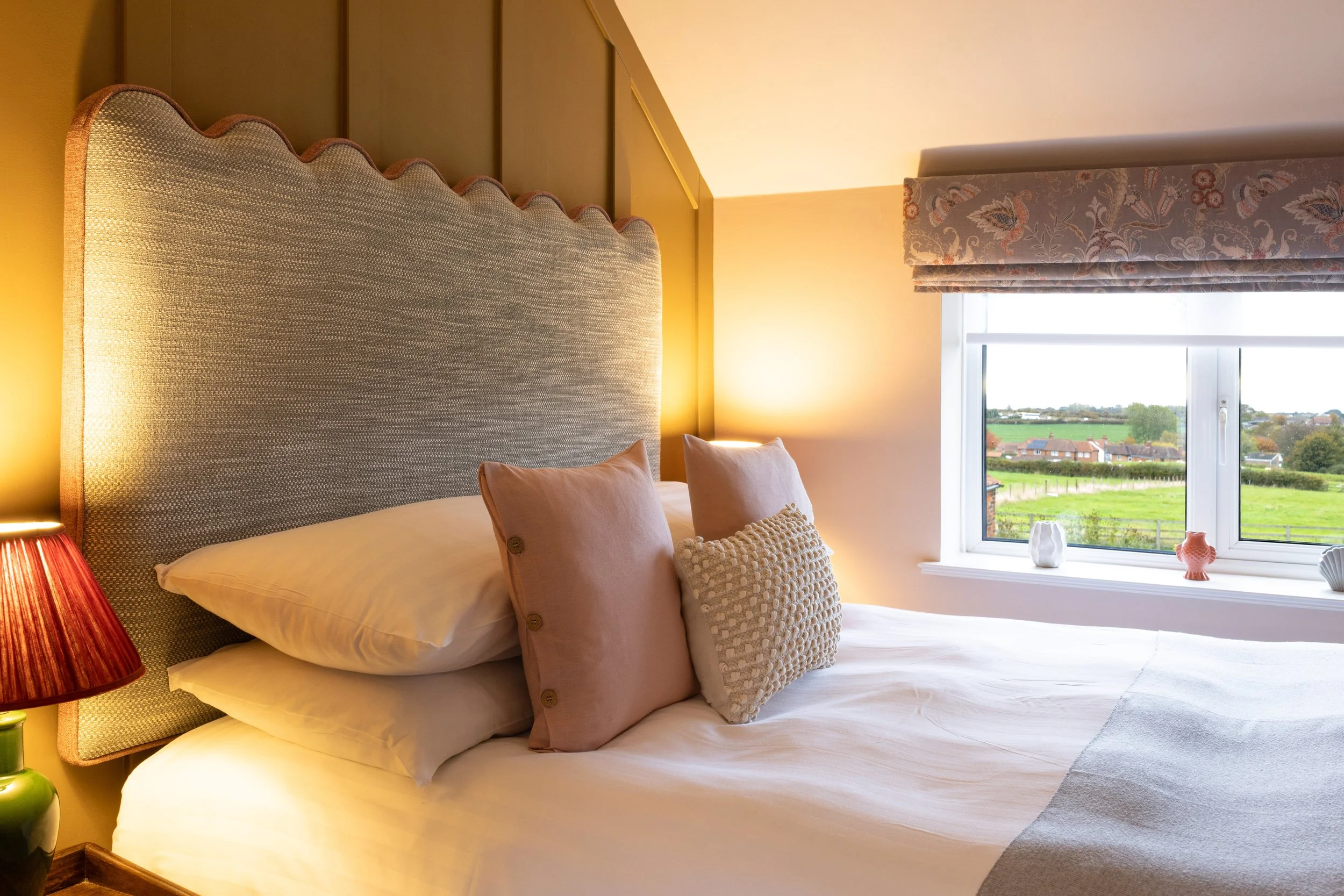 A neatly made bed with a tall, upholstered headboard, two pink pillows, and a textured beige pillow. There is a bedside lamp with a red lampshade, and a window with a floral valance overlooking a green landscape with houses and fields.