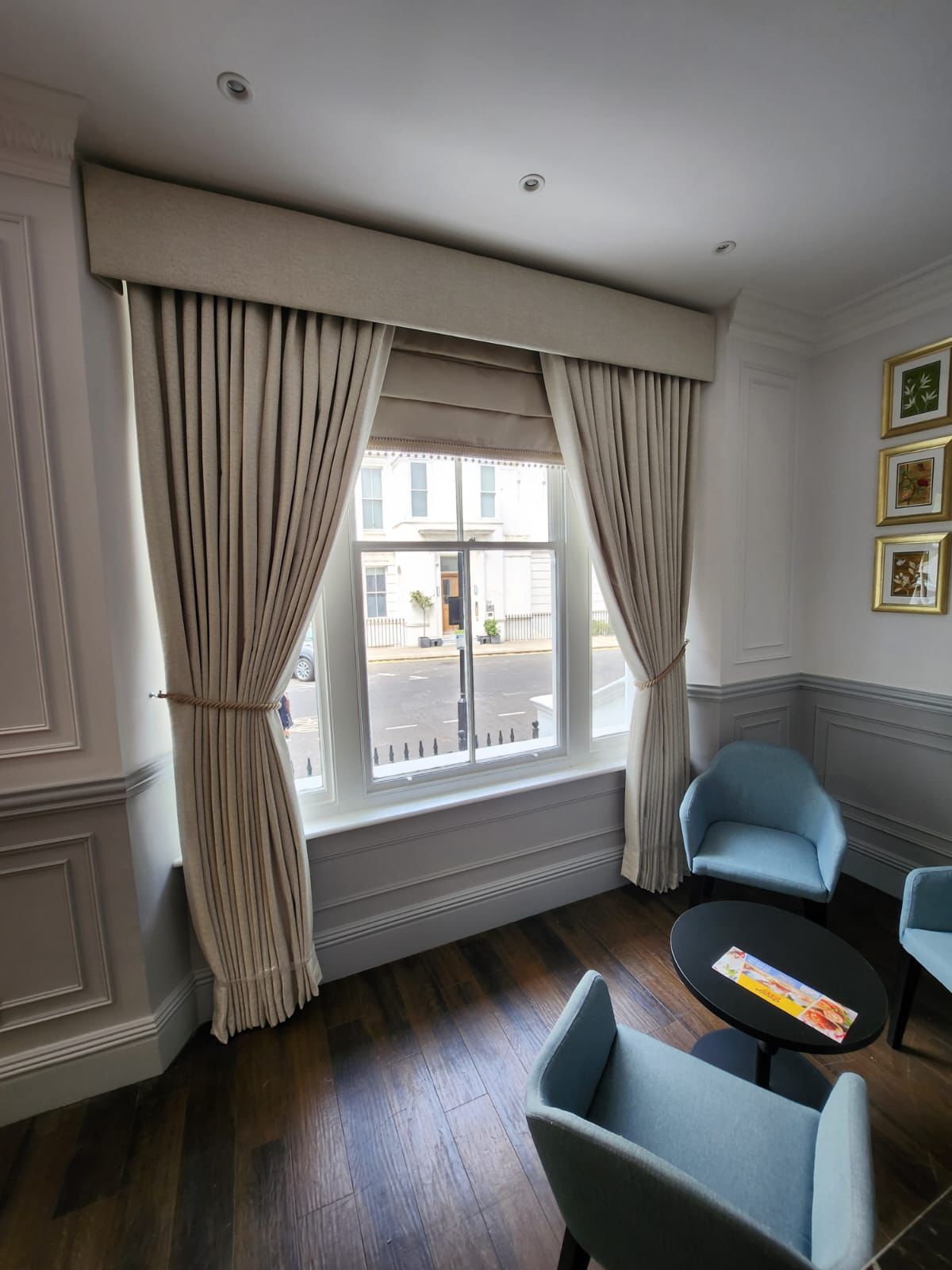 A room with beige curtains framing a window, light blue armchairs, a black round table with a brochure or magazine, white paneled walls with framed artwork, and a dark wooden floor.
