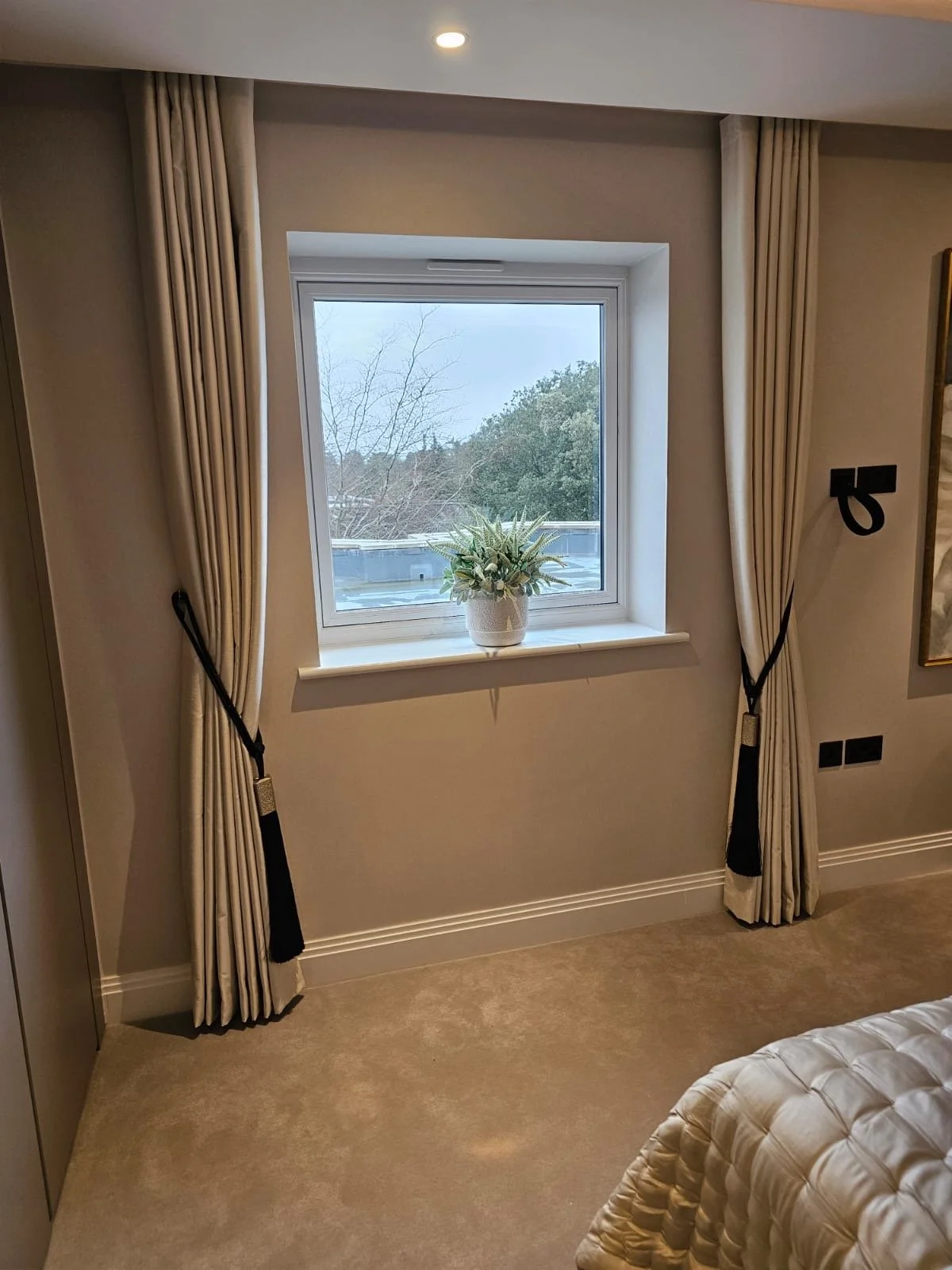 A bedroom window with beige curtains tied back and a white ceramic planter with greenery on the windowsill. Outside, there are trees and a cloudy sky.