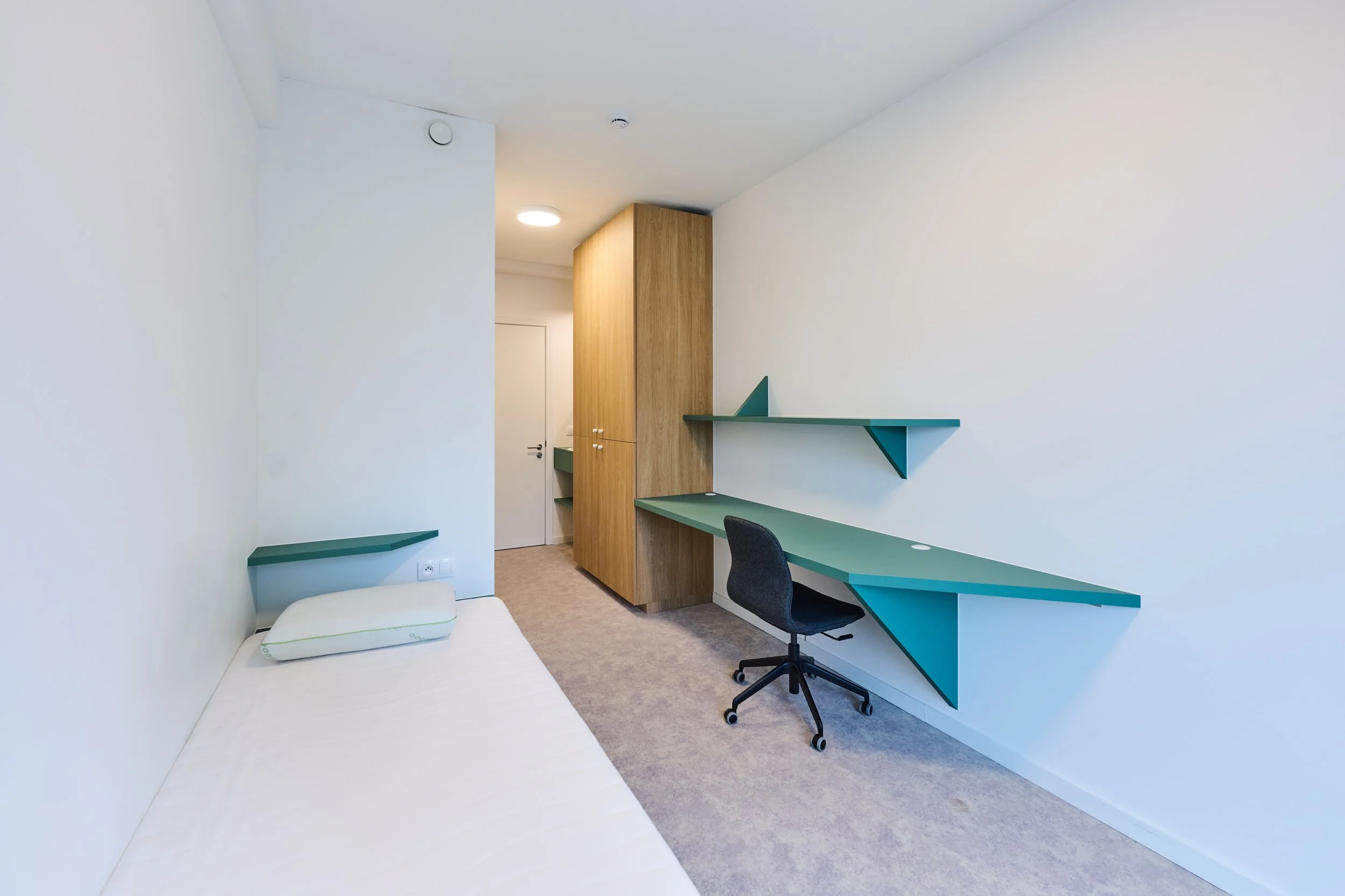 Minimalist bedroom with single bed, green floating desk, and black office chair in white-walled room with wooden storage cabinet.