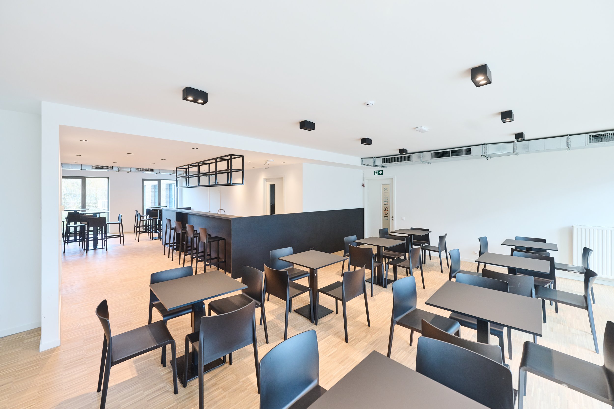 Empty modern dining area with black tables and chairs, bright lighting, and a small bar with barstools.