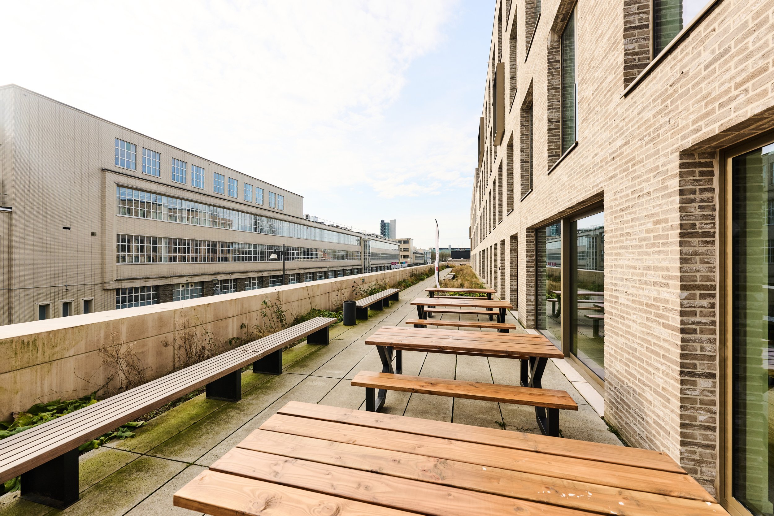Outdoor balcony with wooden tables and benches along the side of a brick building, overlooking a cityscape with large modern buildings.