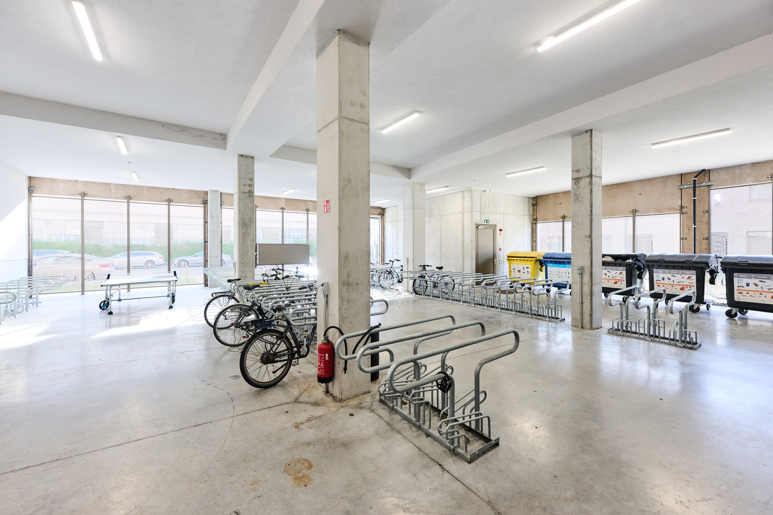 Interior of a bike parking area with several bicycles, bike racks, large trash bins, and windows letting in natural light.