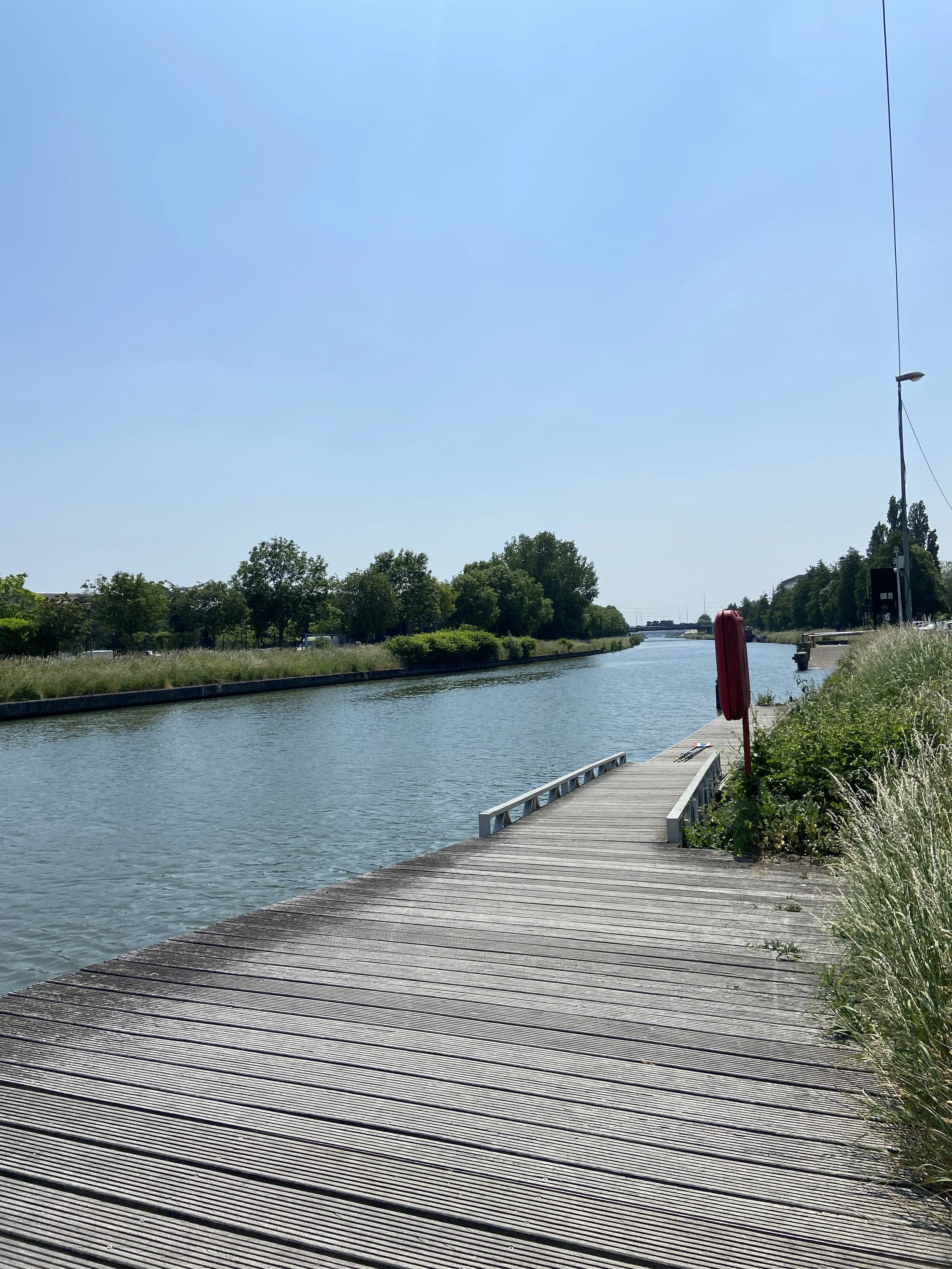 Wooden dock extending over a waterway with greenery and trees on both sides under a clear blue sky.