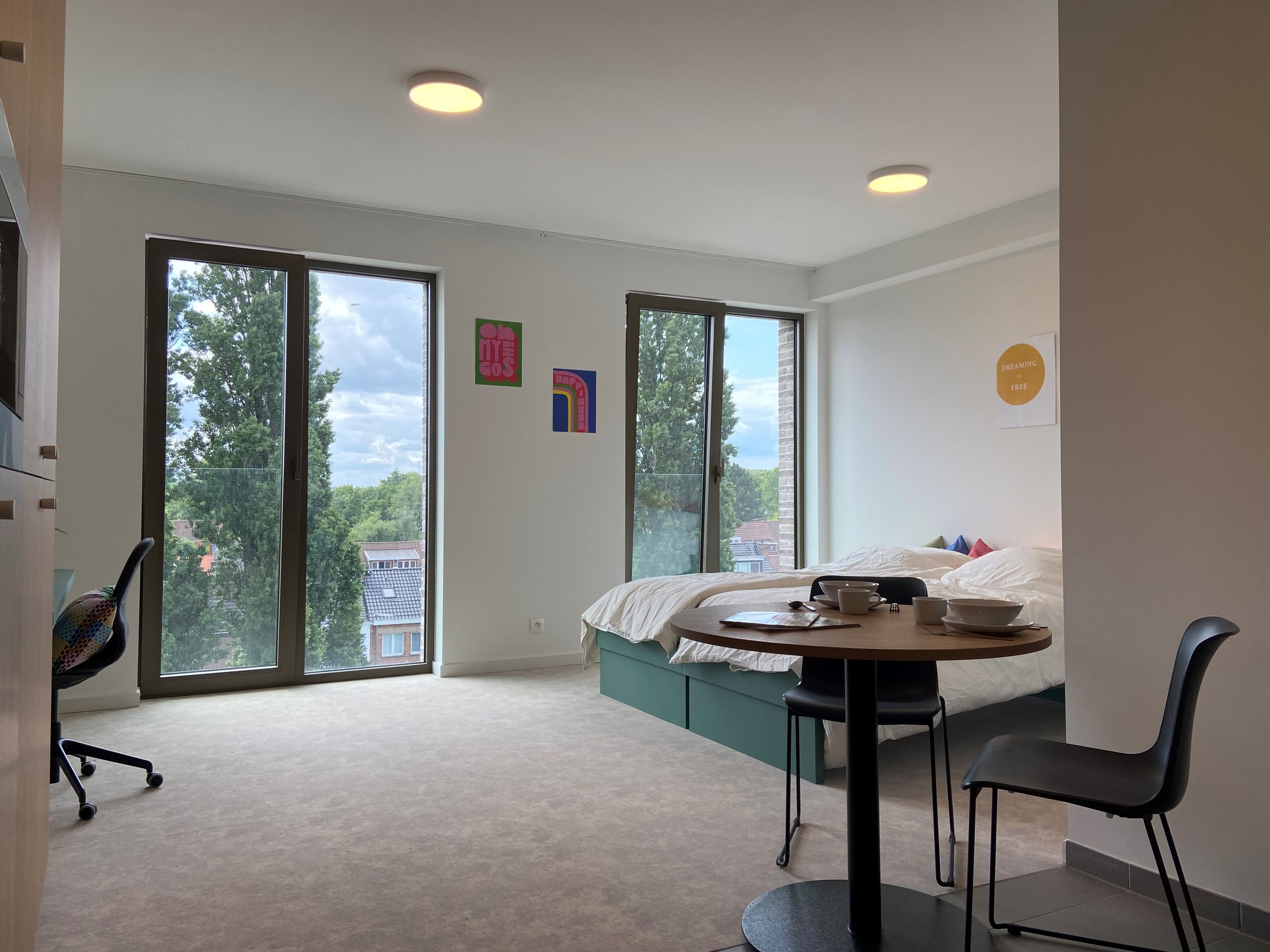 Bright, modern apartment room with large windows, a bed with colorful pillows, a small round dining table with dishes, and a chair. Artwork on the walls and a view of trees and neighborhood rooftops outside.