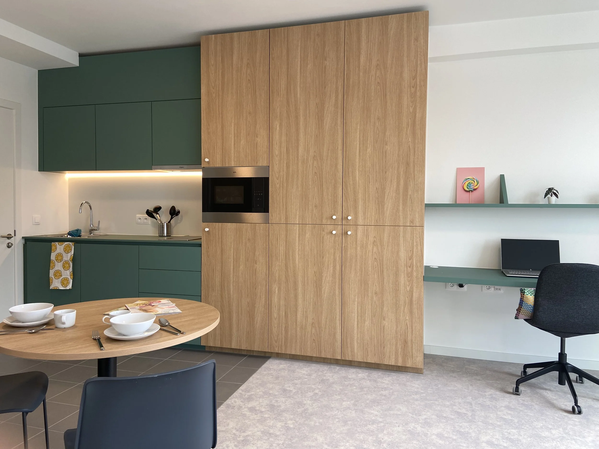 Modern kitchen with green cabinets, wooden storage unit, small dining table set for breakfast, and a black office chair by a built-in desk with a laptop, colorful decorations on a shelf, and a gray rug.