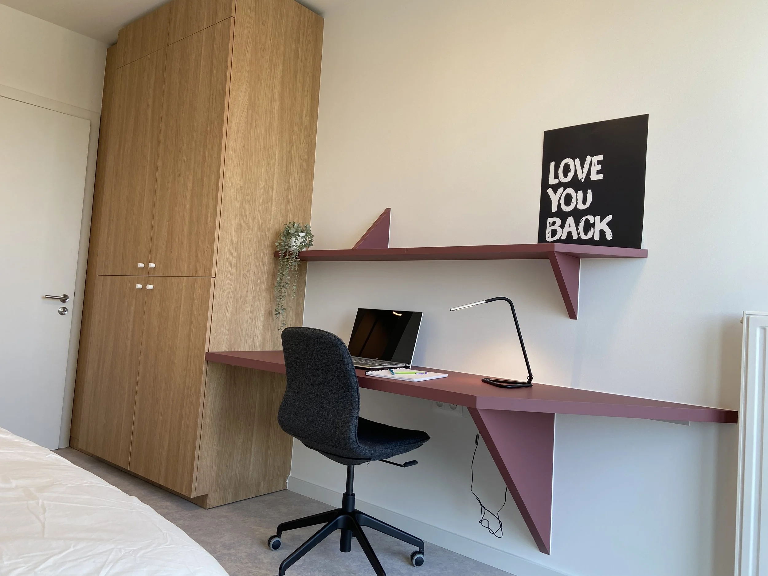 Modern office corner with a black desk lamp, a laptop, and a black office chair. A wooden wardrobe and a wall-mounted mauve-colored desk with an angled design. On the desk is a small plant and a black signboard with white text "LOVE YOU BACK." Above 