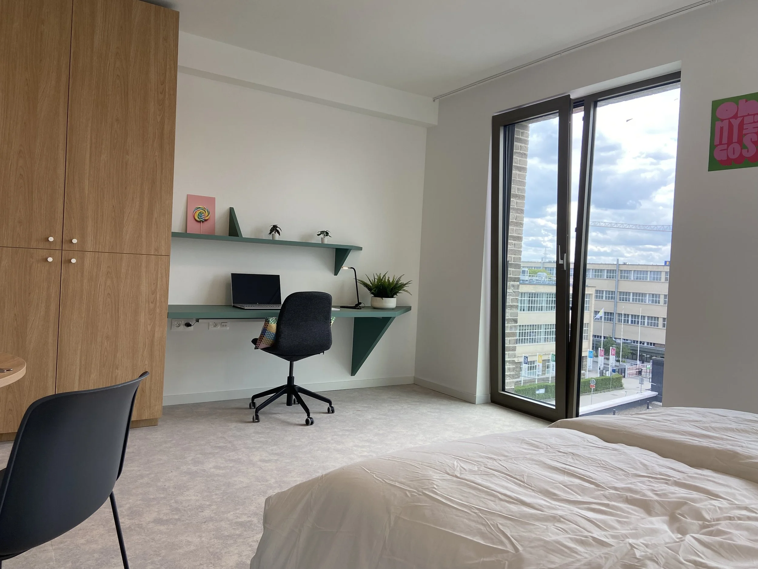 Modern hotel room with a work desk, black office chair, partial view of beds, large glass window with city view, and decorative wall art.