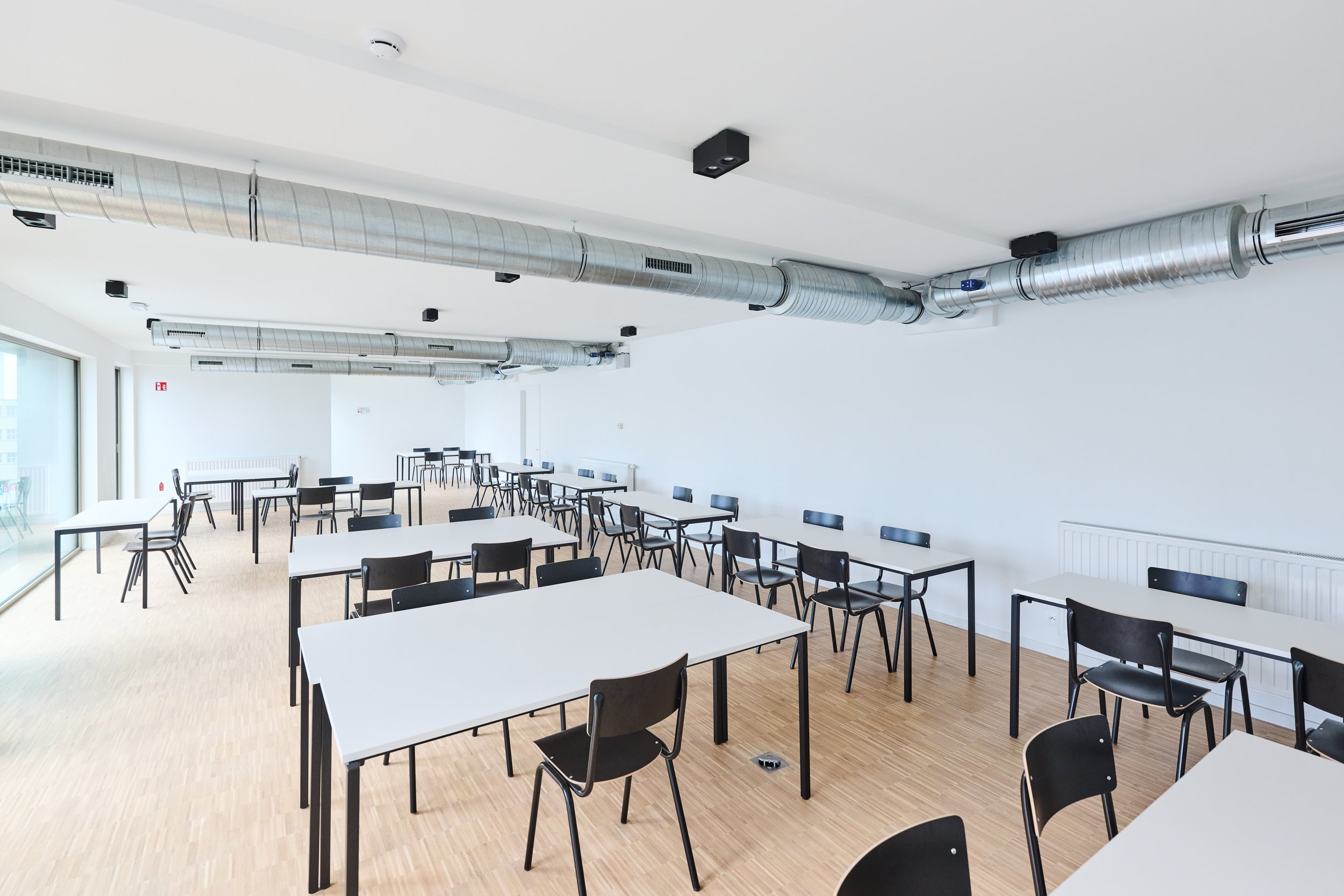 Empty classroom or conference room with white tables and black chairs, large windows, light wood flooring, white walls, and exposed metallic air ducts on the ceiling.