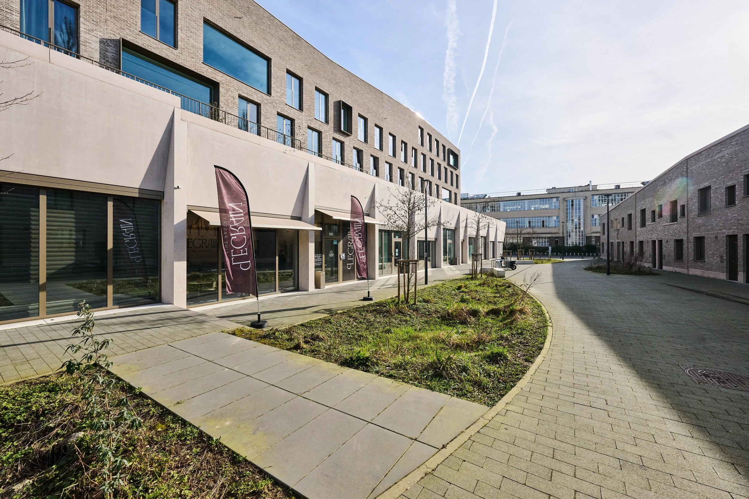 Modern urban courtyard with buildings, shops, leafless trees, and a paved pathway, sunny with clear sky and contrails.