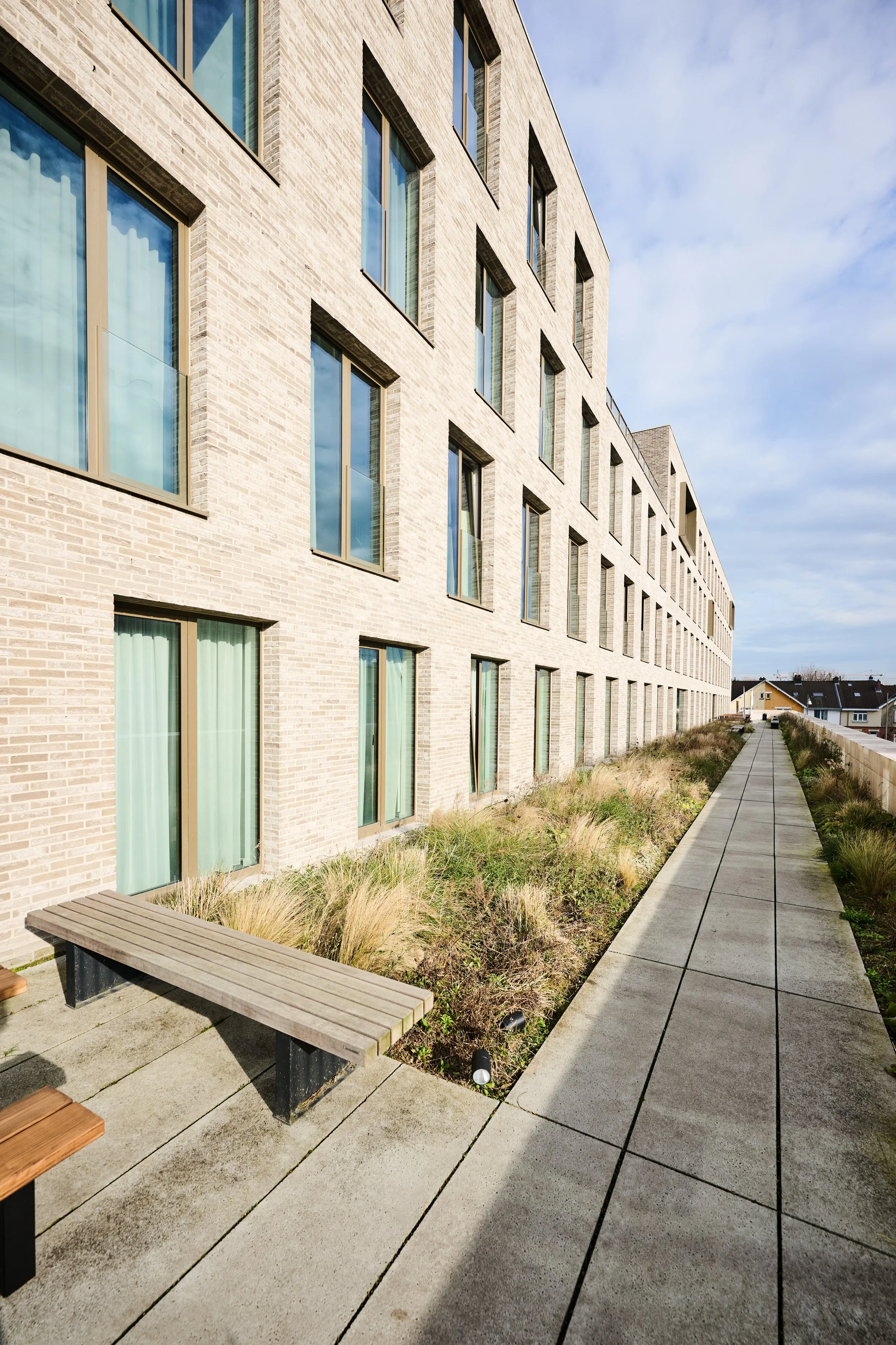 A modern multi-story apartment building with numerous large windows, next to a concrete walkway, benches, and landscaped outdoor space with grass and bushes, under a partly cloudy sky.