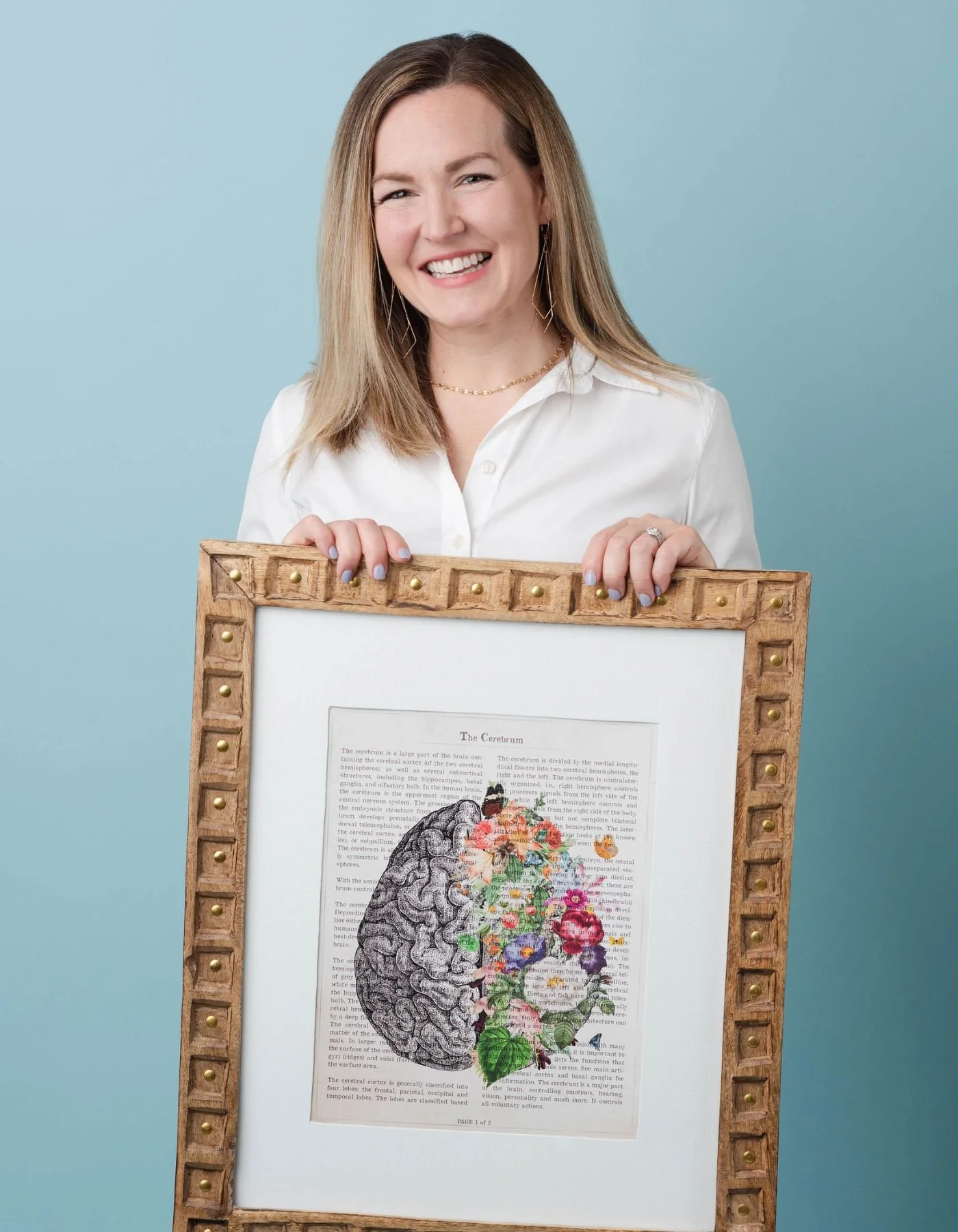 Integrative psychiatrist Dr. Stacey Elliott, wearing a white shirt, holding a framed illustration of a brain with colorful flowers on the right side and text in the background.