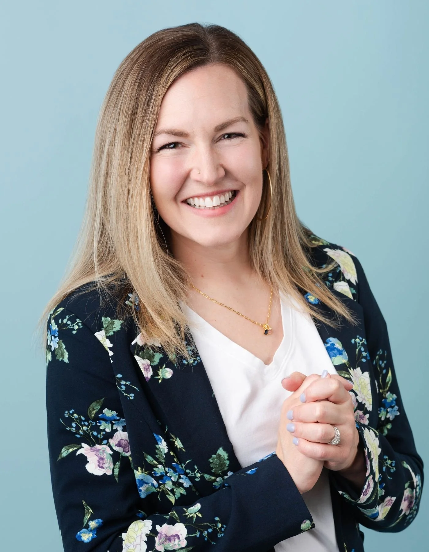 Integrative psychiatrist Dr. Stacey Elliott, smiling, wearing a floral blazer over a white top, gold necklace, and ring, with hands clasped in front, against a light blue background.