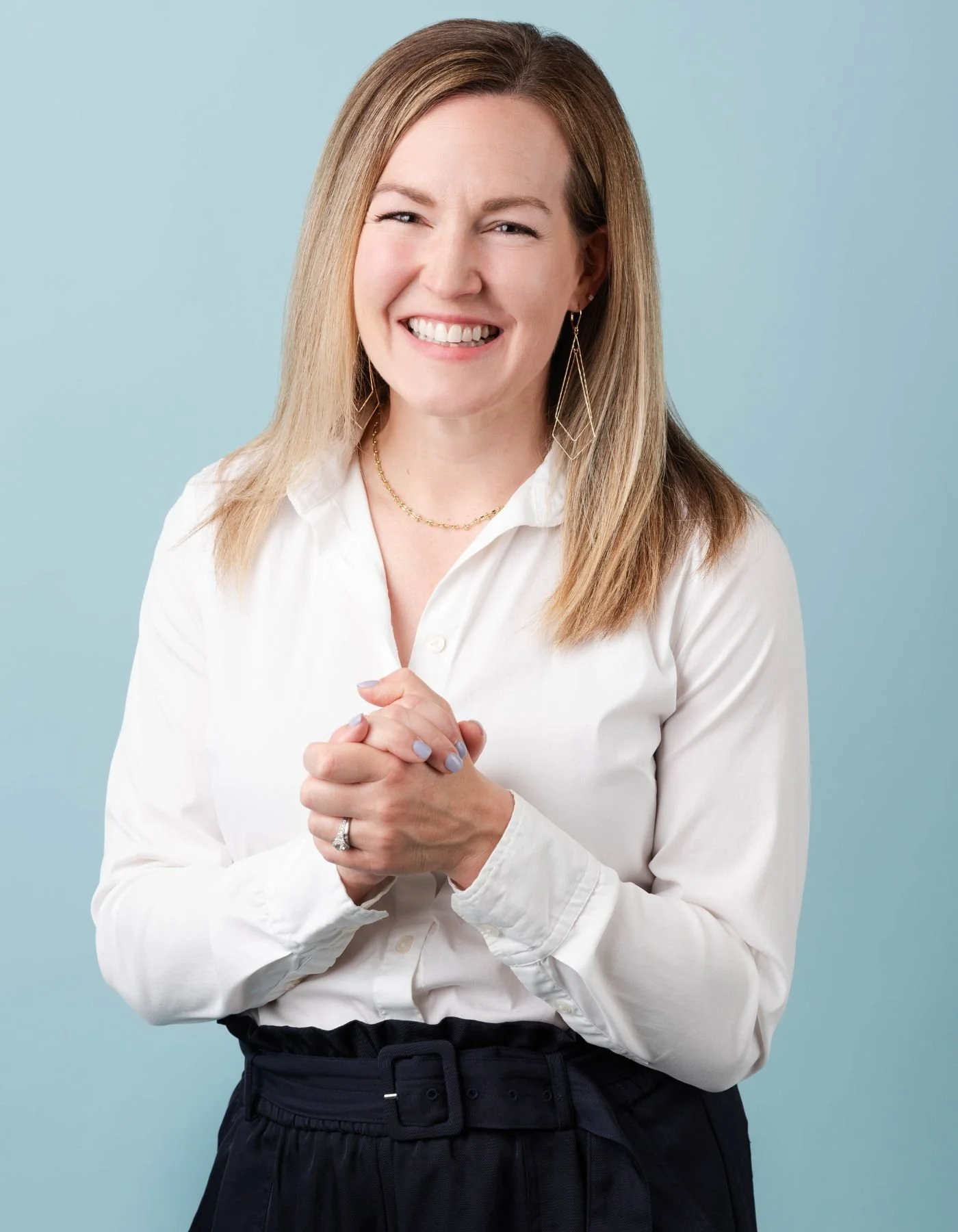 Integrative psychiatrist Dr. Stacey Elliott, wearing a long-sleeve white blouse and high-waisted blue pants clasping her hands and smiling warmly at the camera.
