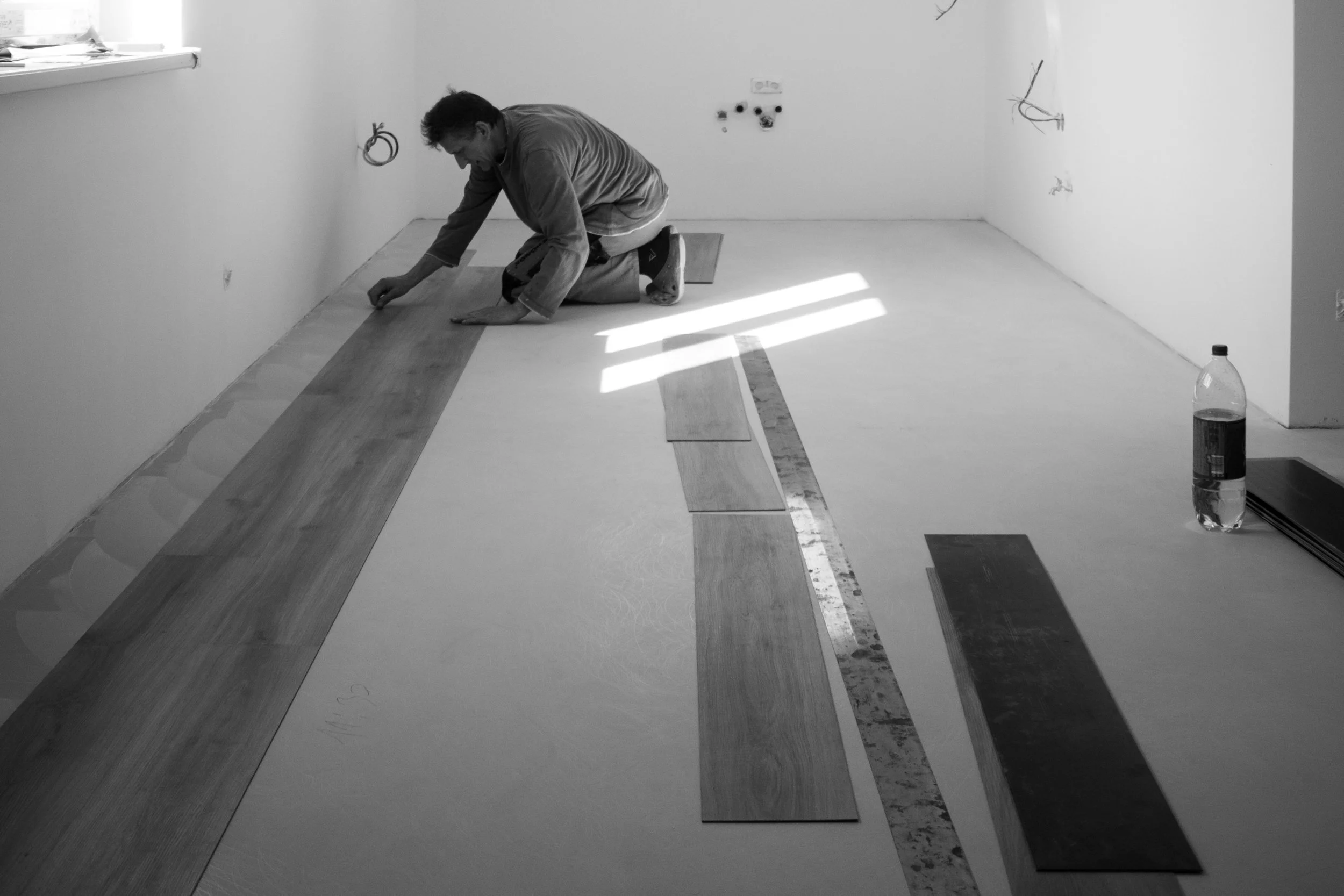 A person kneeling on the floor laying down wooden floorboards in a room under construction, with unfinished walls and visible wiring.