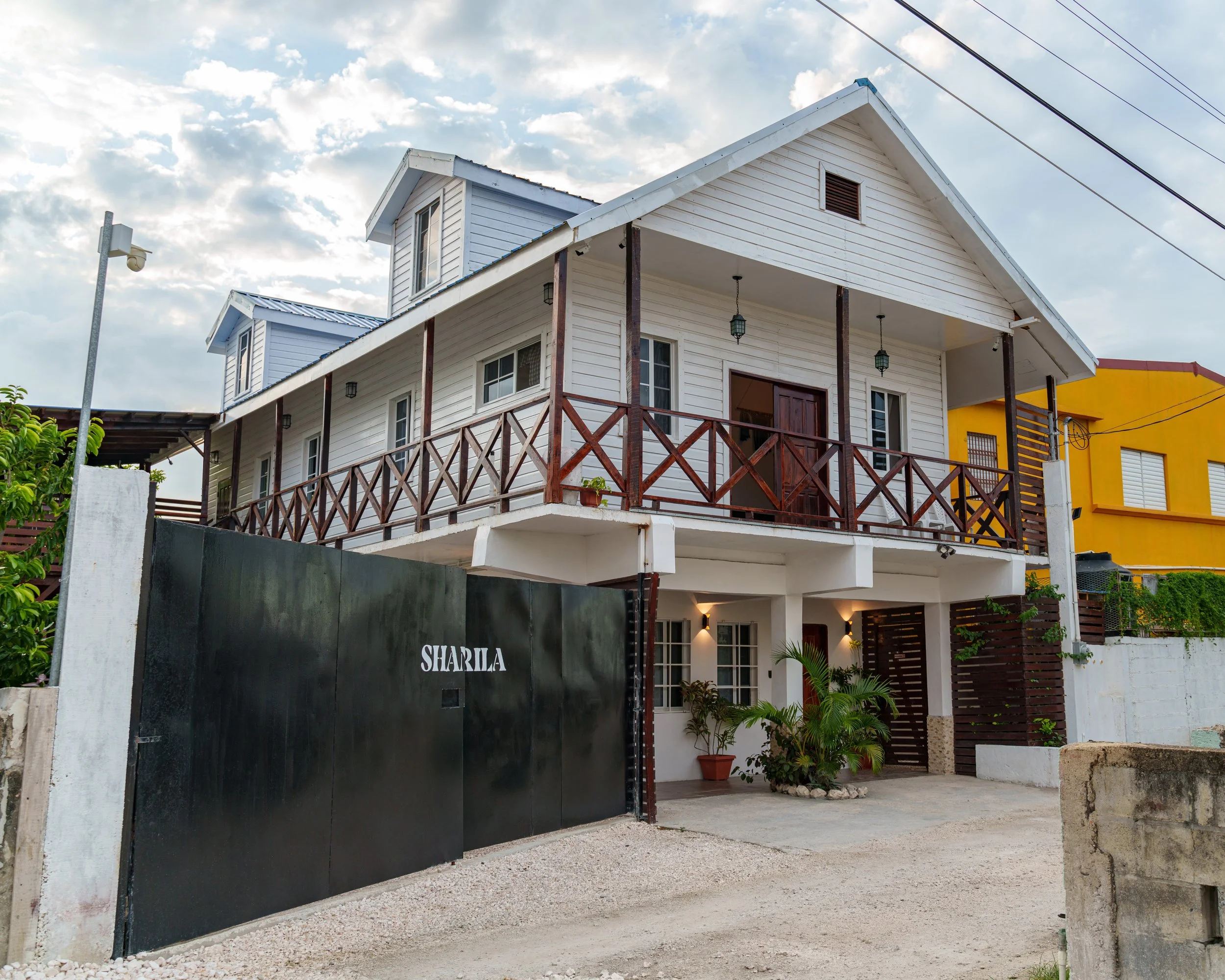 A two-story house with white wooden siding, a covered balcony, and a gated entrance with the name 'SHARIA' on the gate. The house has a gable roof, small dormer windows, and plants near the entrance.