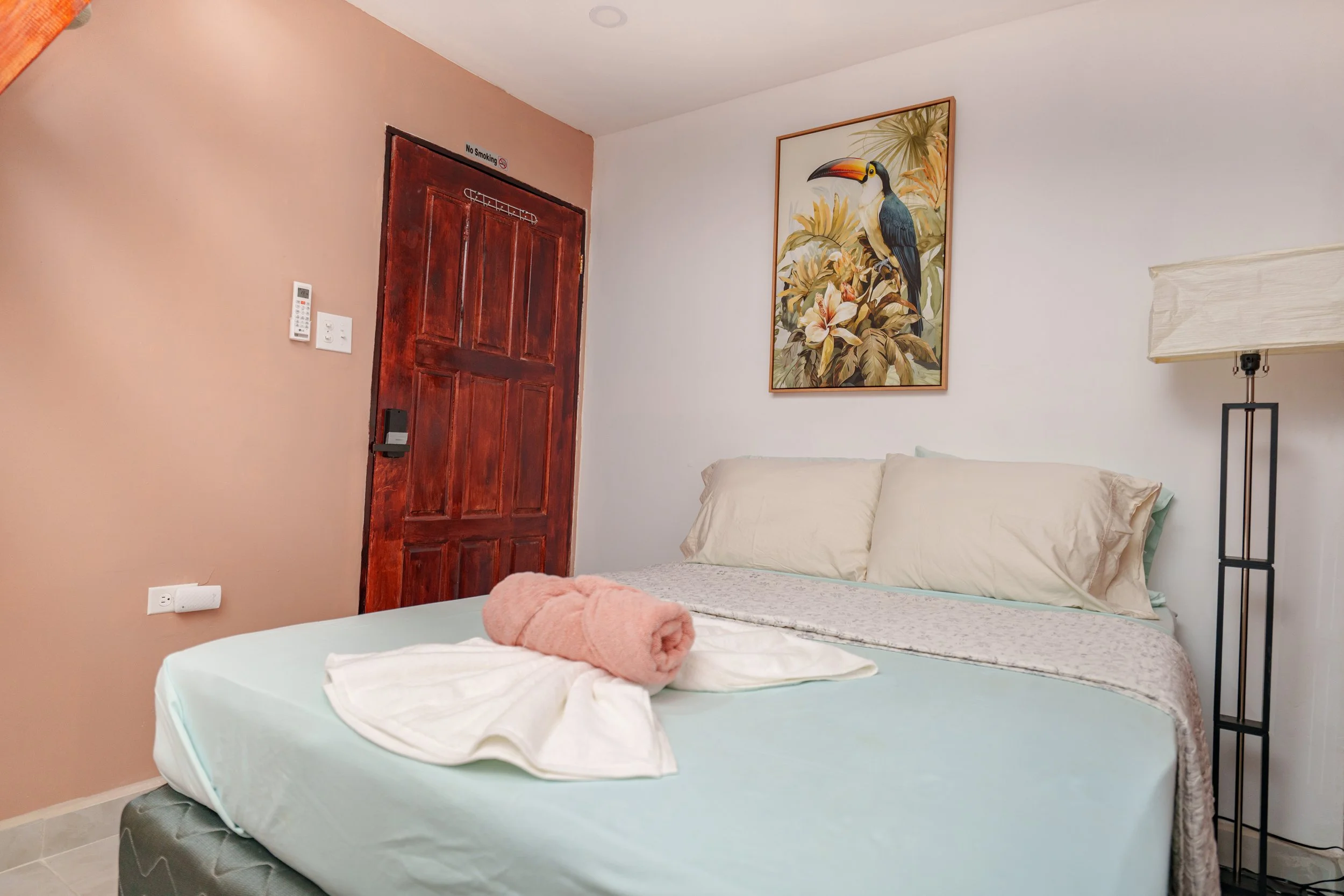 A neatly made bed with beige pillows and a rolled pink towel on top. A framed tropical bird artwork hangs above the bed. A tall floor lamp stands beside the bed. The room has a white and peach-colored wall with a wooden door.