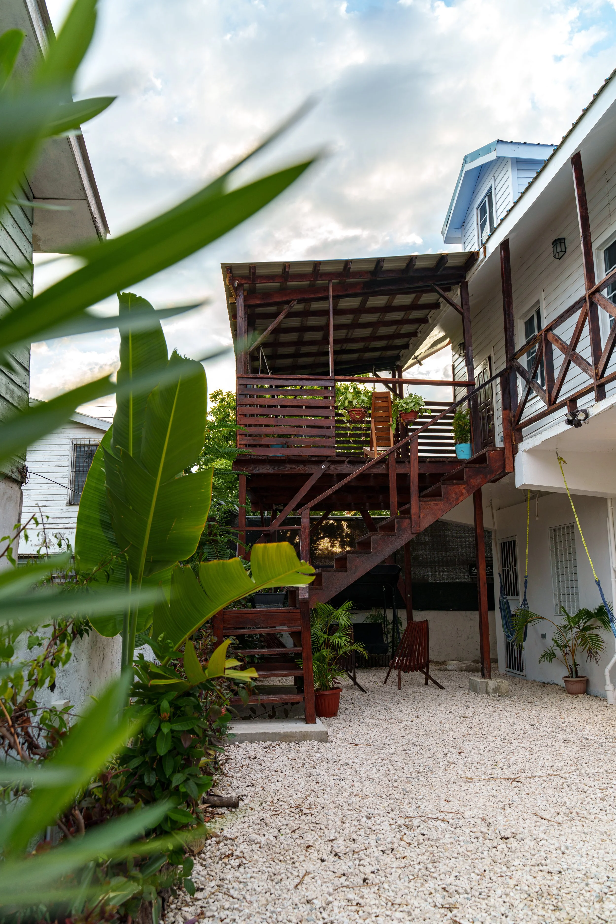 Backyard with gravel ground, potted plants, wooden staircase leading to an upper porch with plants, white house wall, and blue house roof visible in the background under a partly cloudy sky.