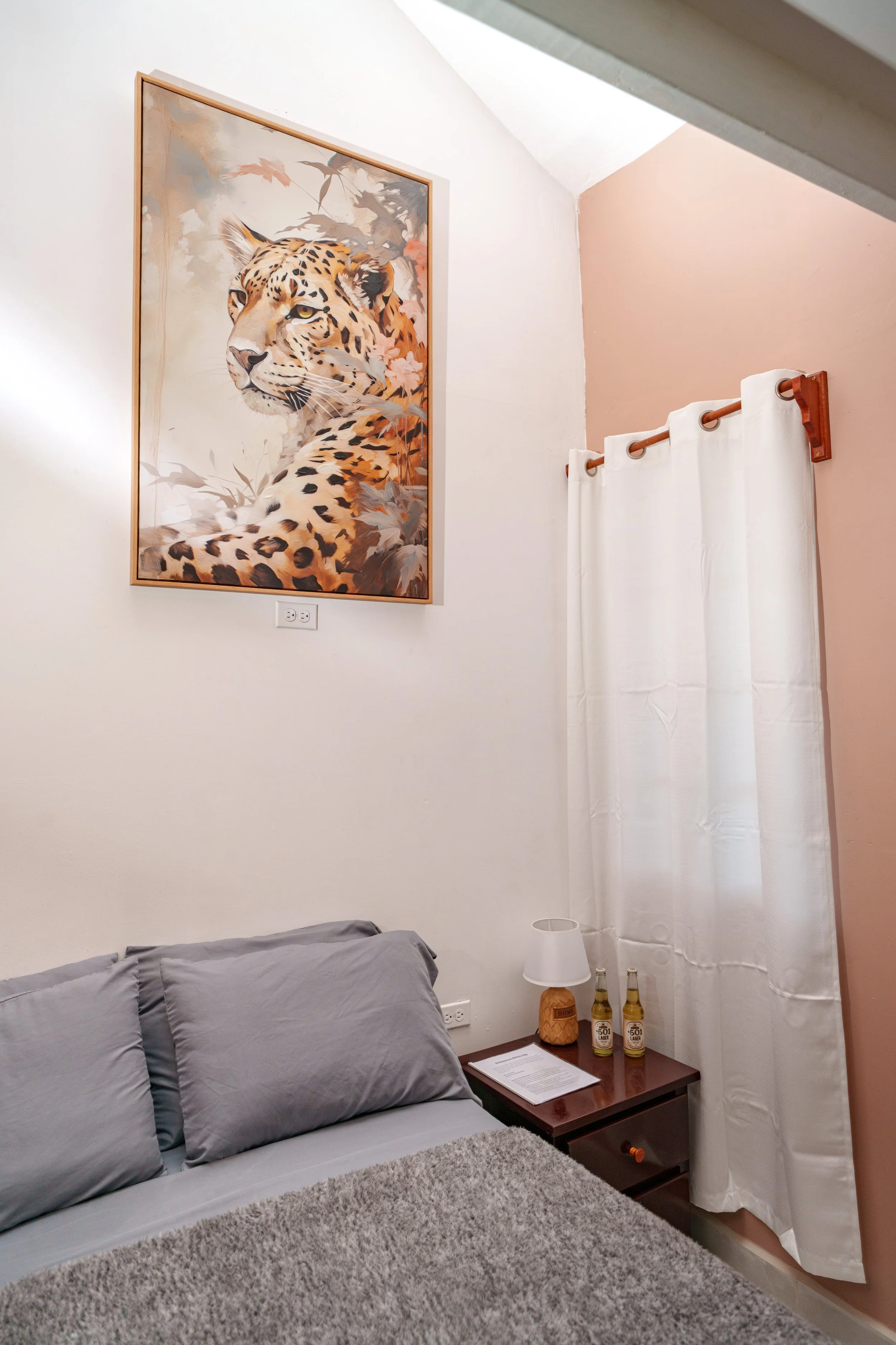 A bedroom corner with a bed, gray pillows, a nightstand with two bottles, a lamp, a paper, a white curtain, and a large painting of a cheetah on the wall.