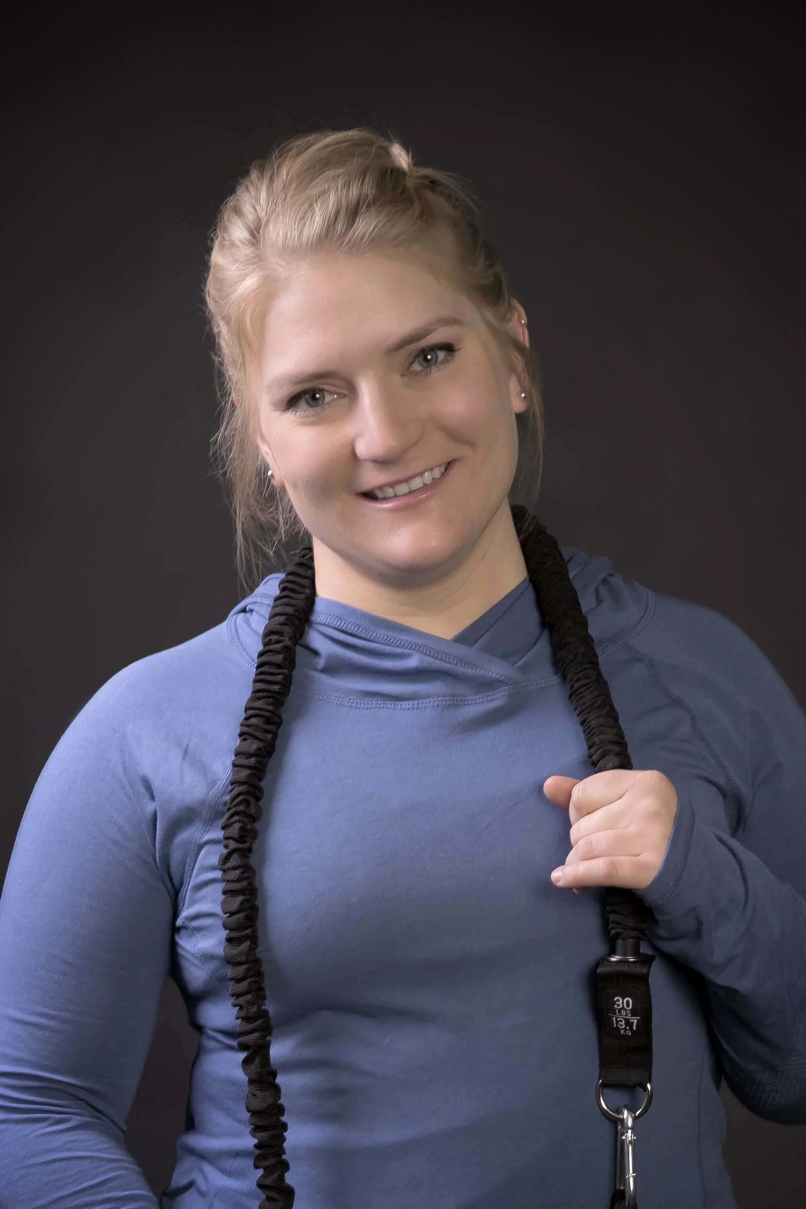 A woman with blonde hair in a braid wearing a blue workout shirt, holding a jump rope over her shoulder, smiling at the camera against a dark background.