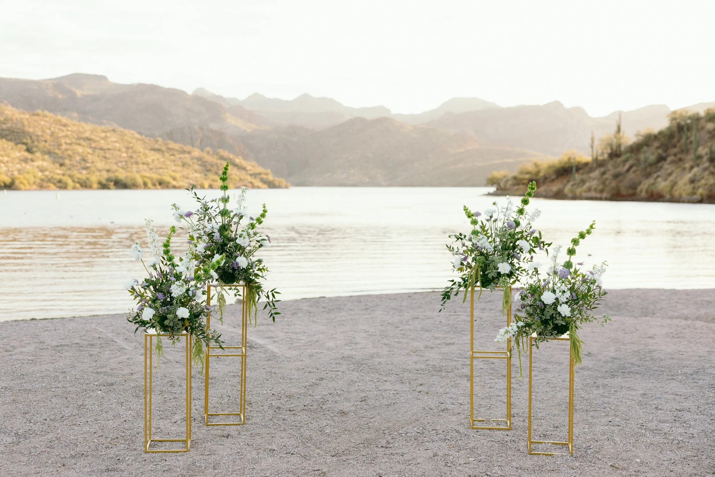 Wedding aisle with four flower arrangements on gold stands by a lake with mountains in the background.