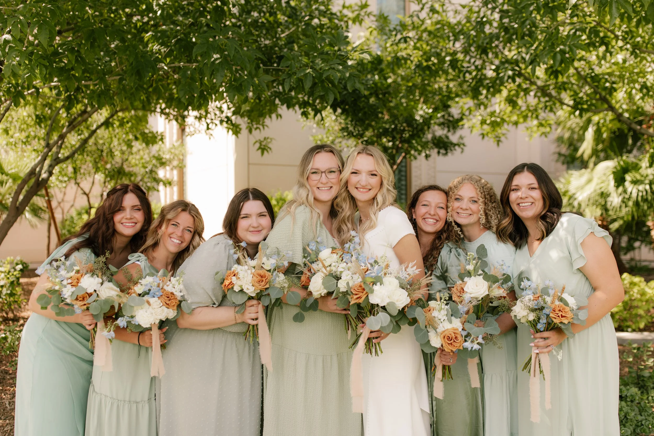 A group of nine women, including a bride and bridesmaids, smiling and holding bouquets of flowers, standing outdoors under tree foliage.
