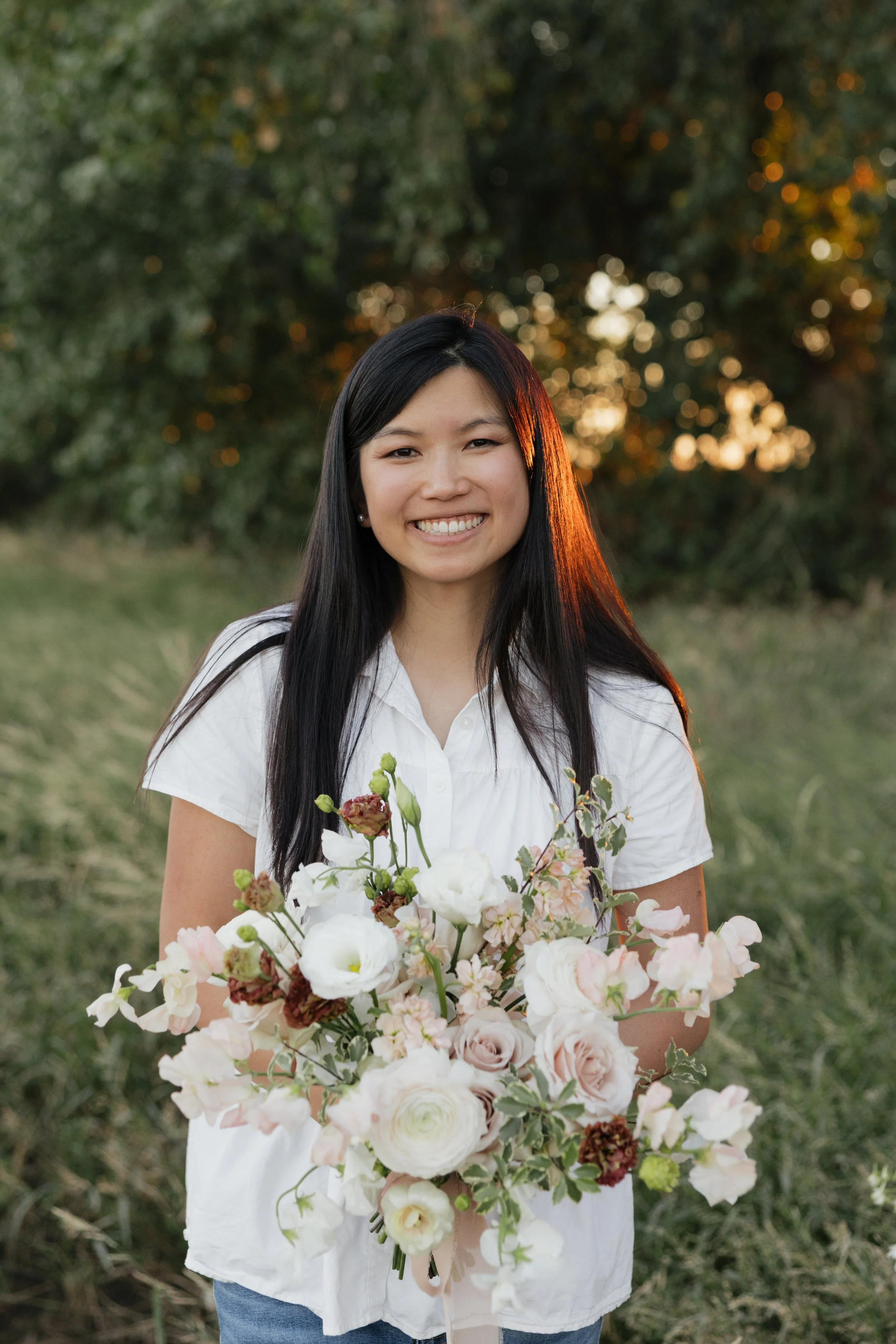 A young woman with long black hair smiling and holding a bouquet of assorted flowers outdoors during sunset.