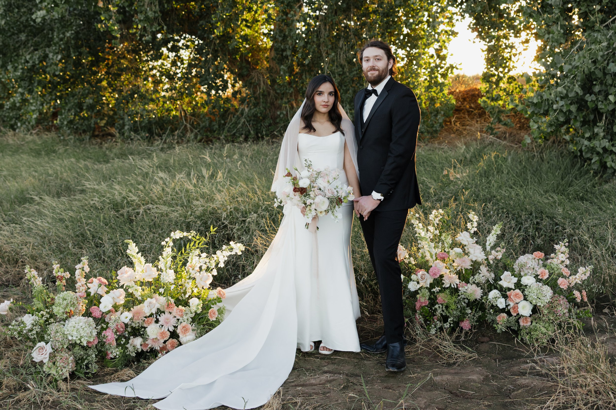 A bride and groom holding hands outdoors during sunset, surrounded by flowers and trees.