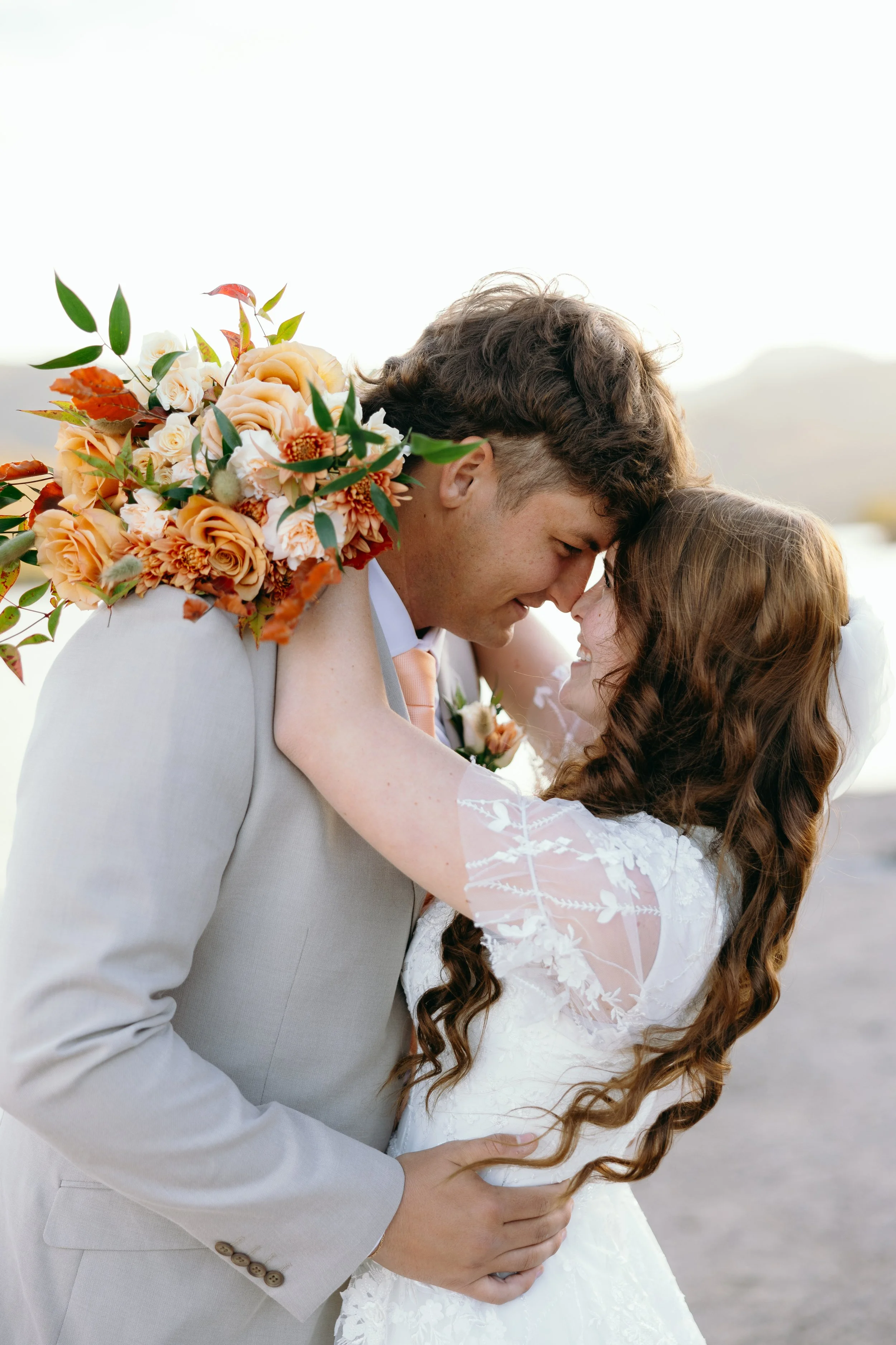 Bride and groom embracing outdoors on their wedding day, smiling and touching foreheads, with a bouquet of flowers and a natural background.