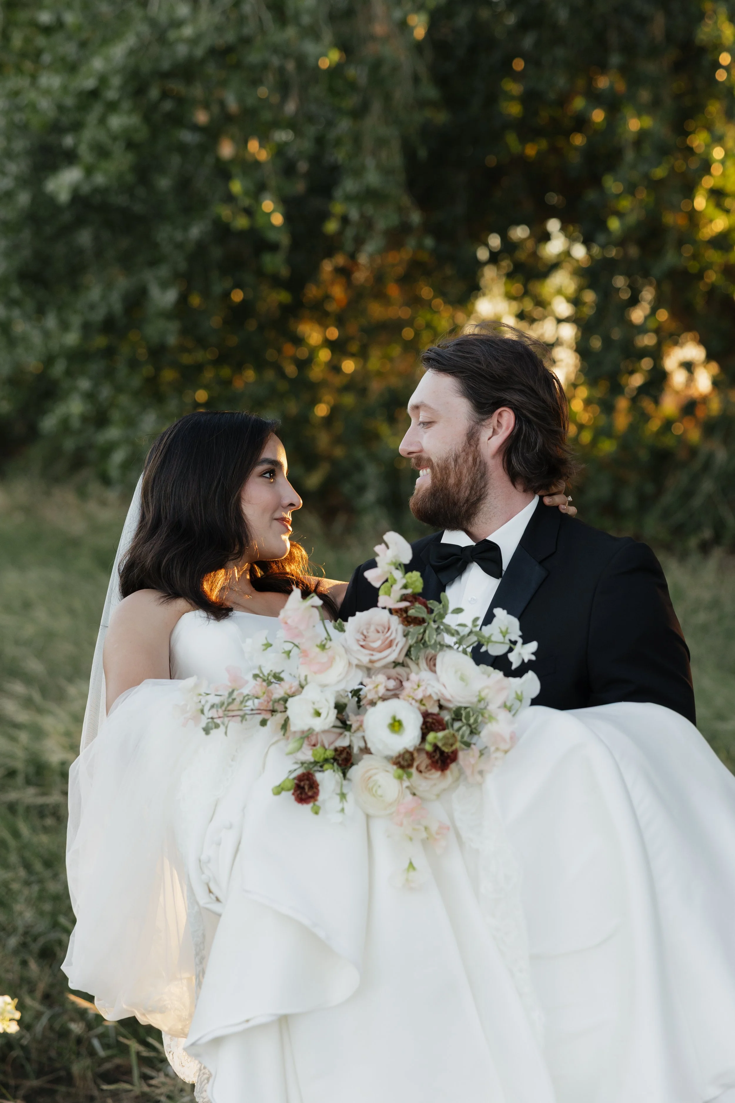A newlywed couple in wedding attire, holding a bouquet of flowers, looking at each other outdoors during sunset.