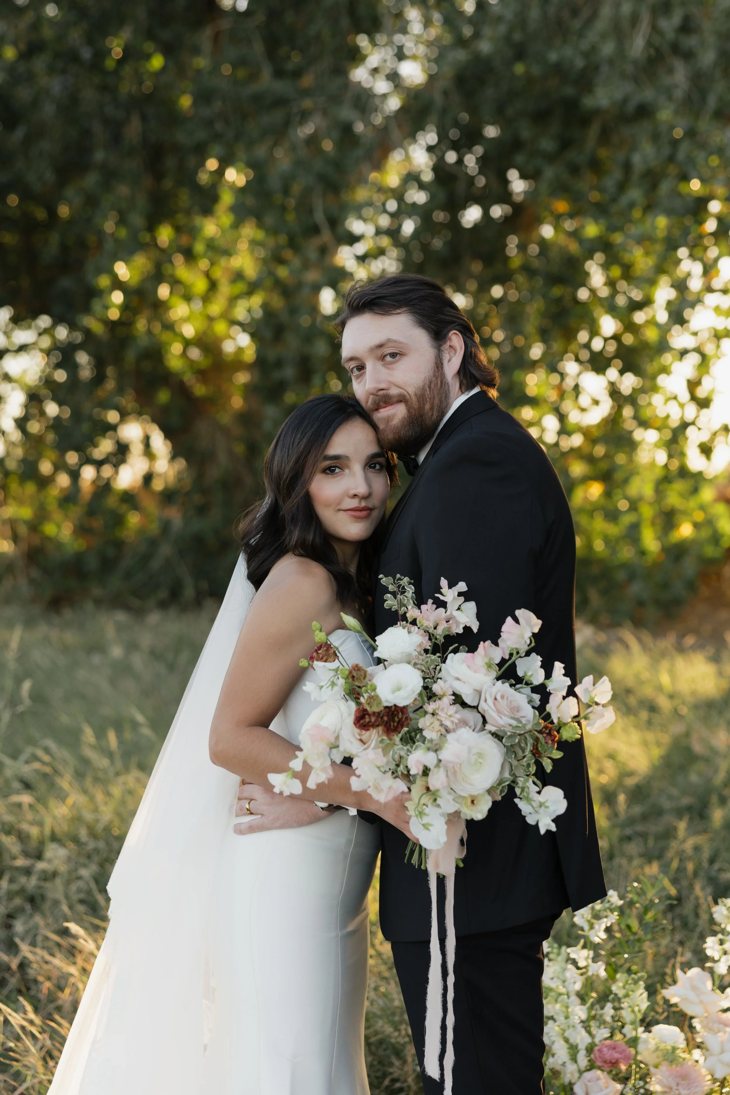 A bride and groom pose outdoors during their wedding, with the bride holding a large bouquet of white and pink flowers, and the groom dressed in a black tuxedo. The background features greenery and sunlight filtering through trees.