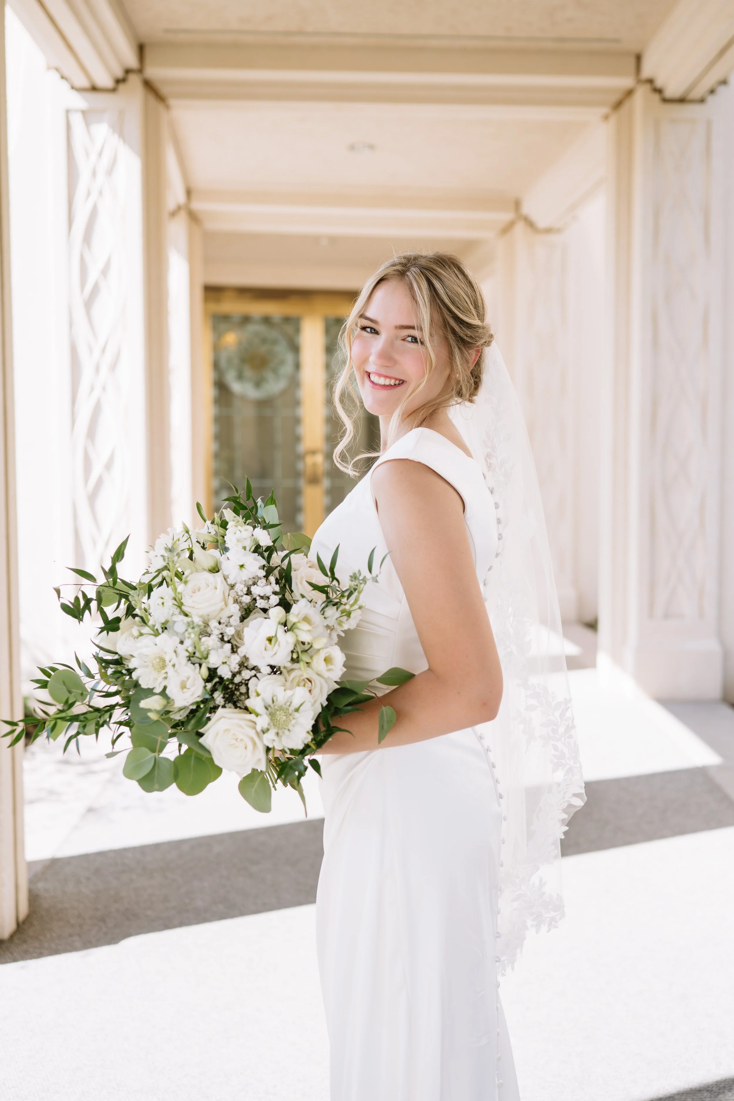 A bride in a white wedding dress holding a bouquet of white flowers and greenery, smiling and looking at the camera, standing in a decorated indoor setting.
