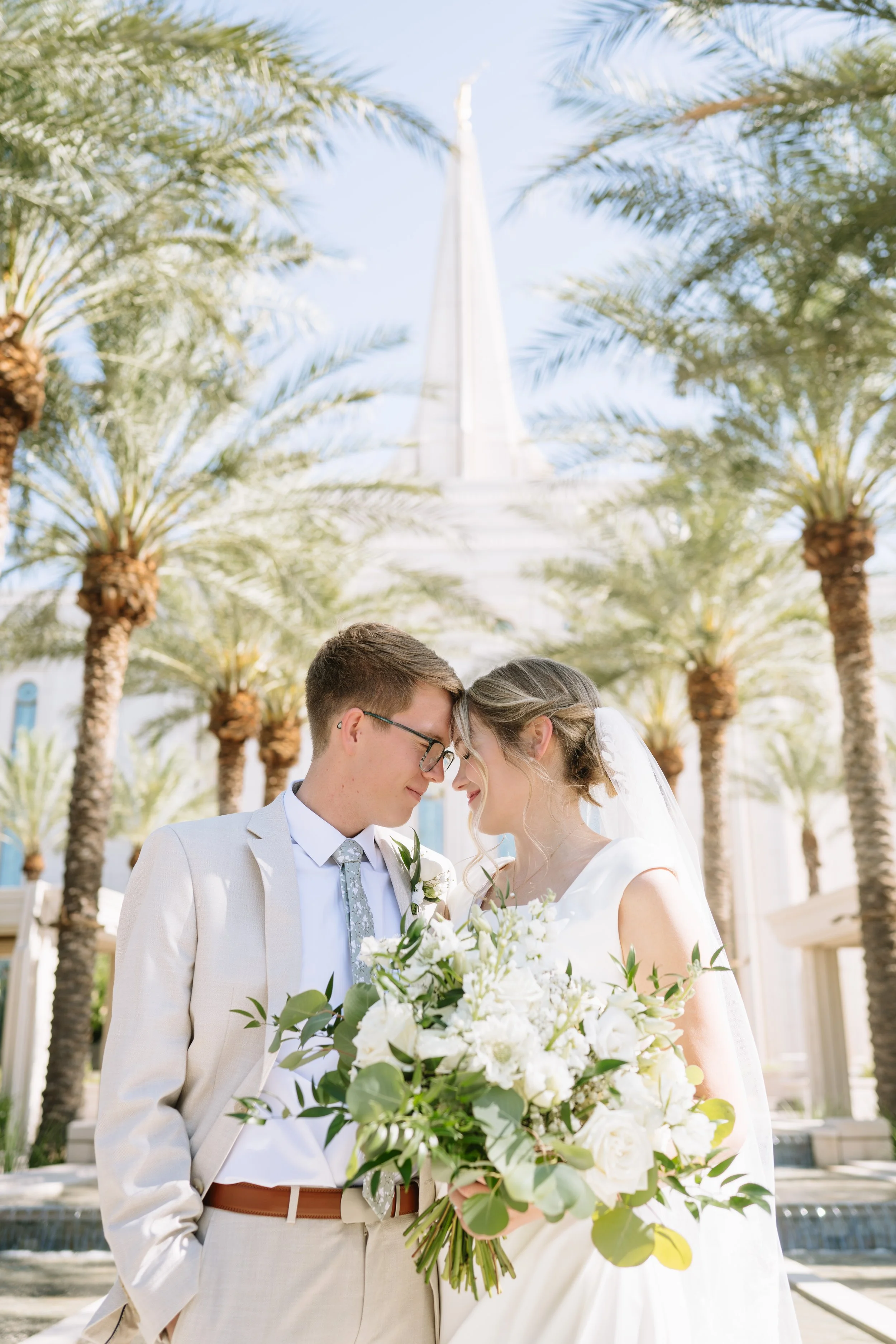 A bride and groom face each other with foreheads touching, holding a large bouquet of white flowers, standing outdoors with palm trees and a tall white building in the background.