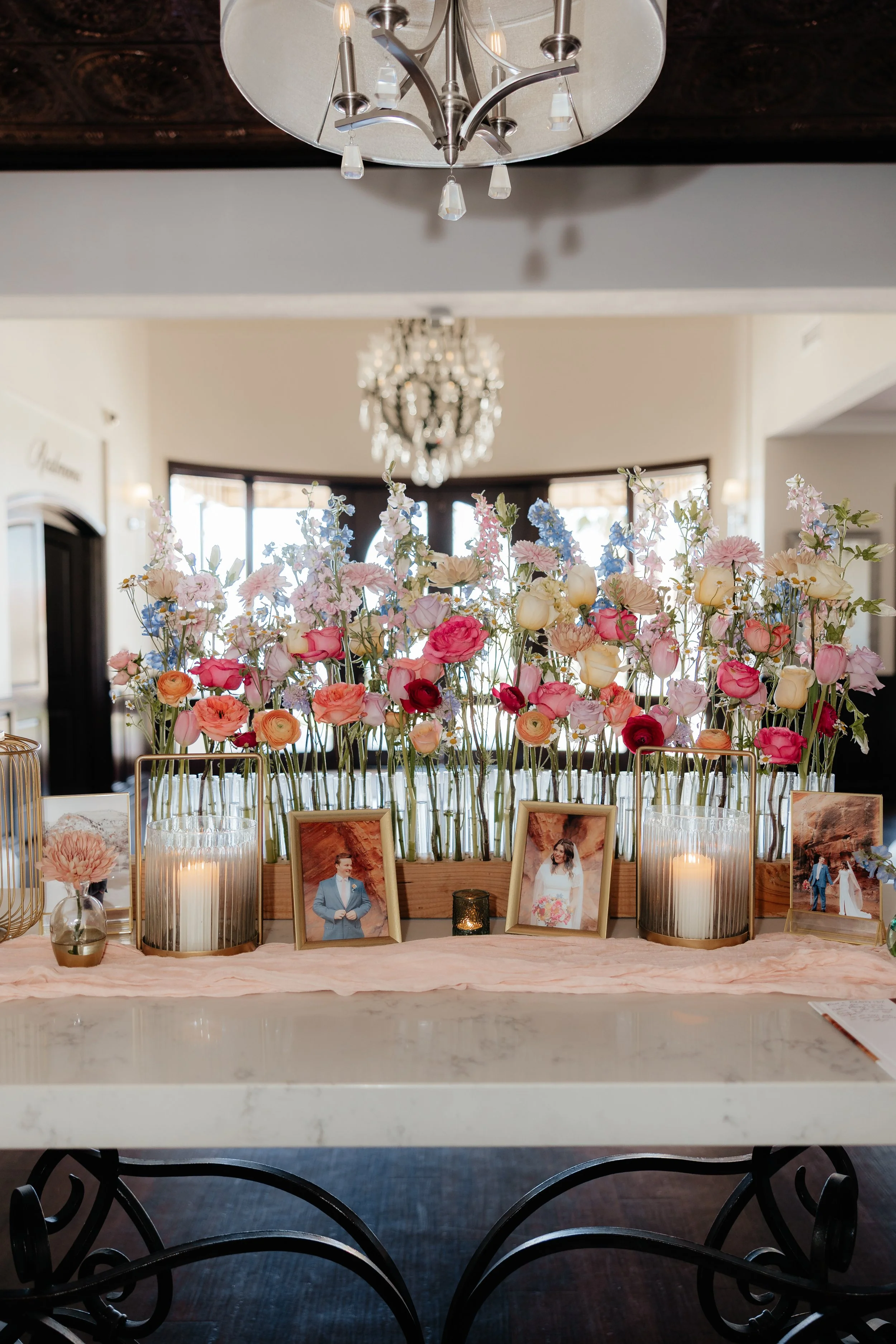 A floral display with pink, white, and peach flowers in glass and wooden containers, framed photos, and candles on a table, in a bright indoor setting.