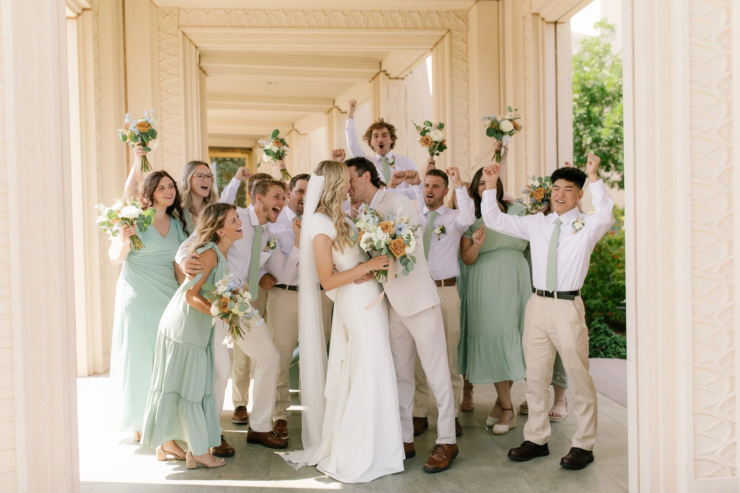A wedding celebration with the bride and groom kissing in the center, surrounded by guests cheering, holding bouquets, and raising fists in celebration, inside a decorated venue with natural light.