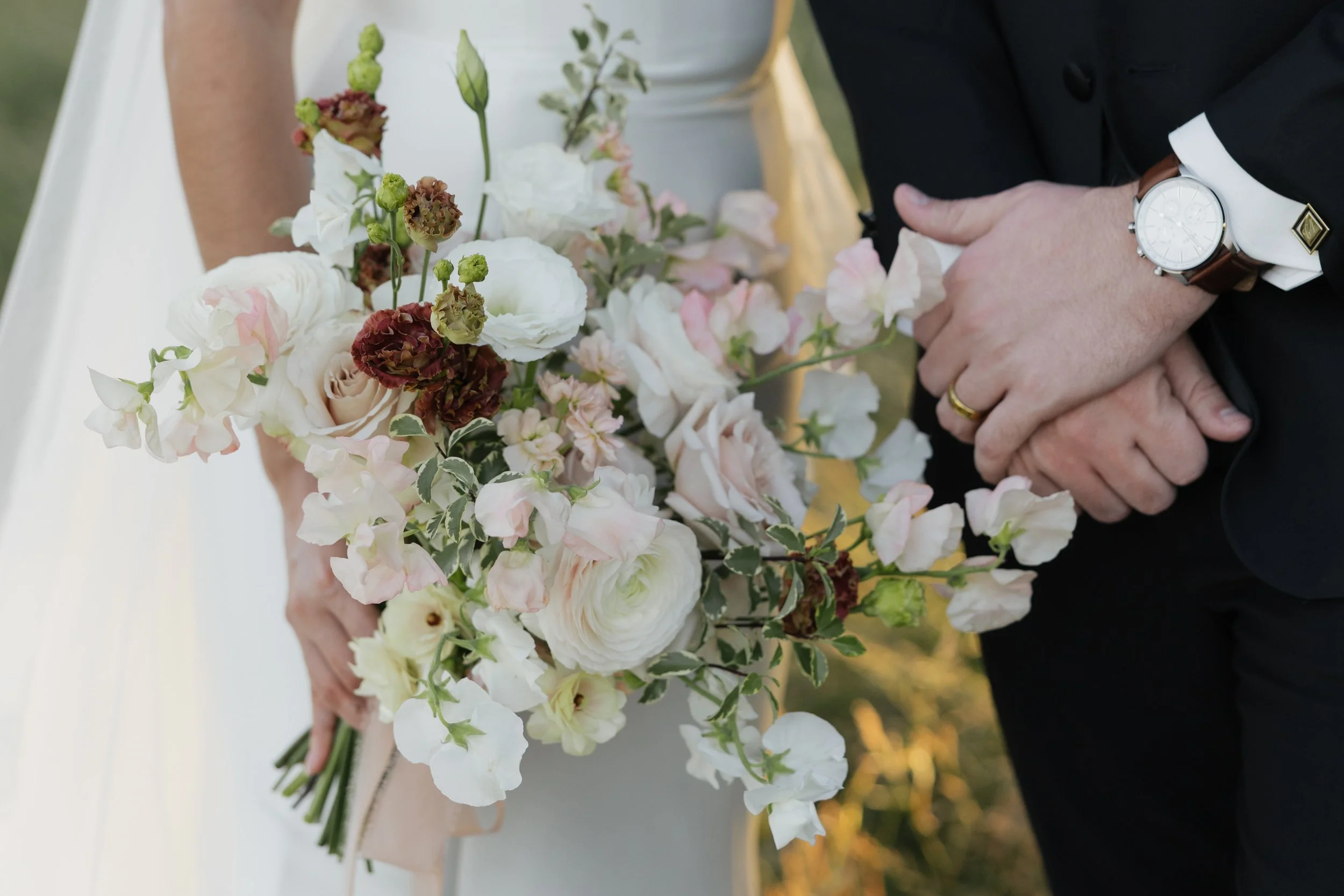 Close-up of a wedding bouquet with white, blush pink, and dark red flowers, held by a bride, with a groom in a black suit and watch standing nearby.