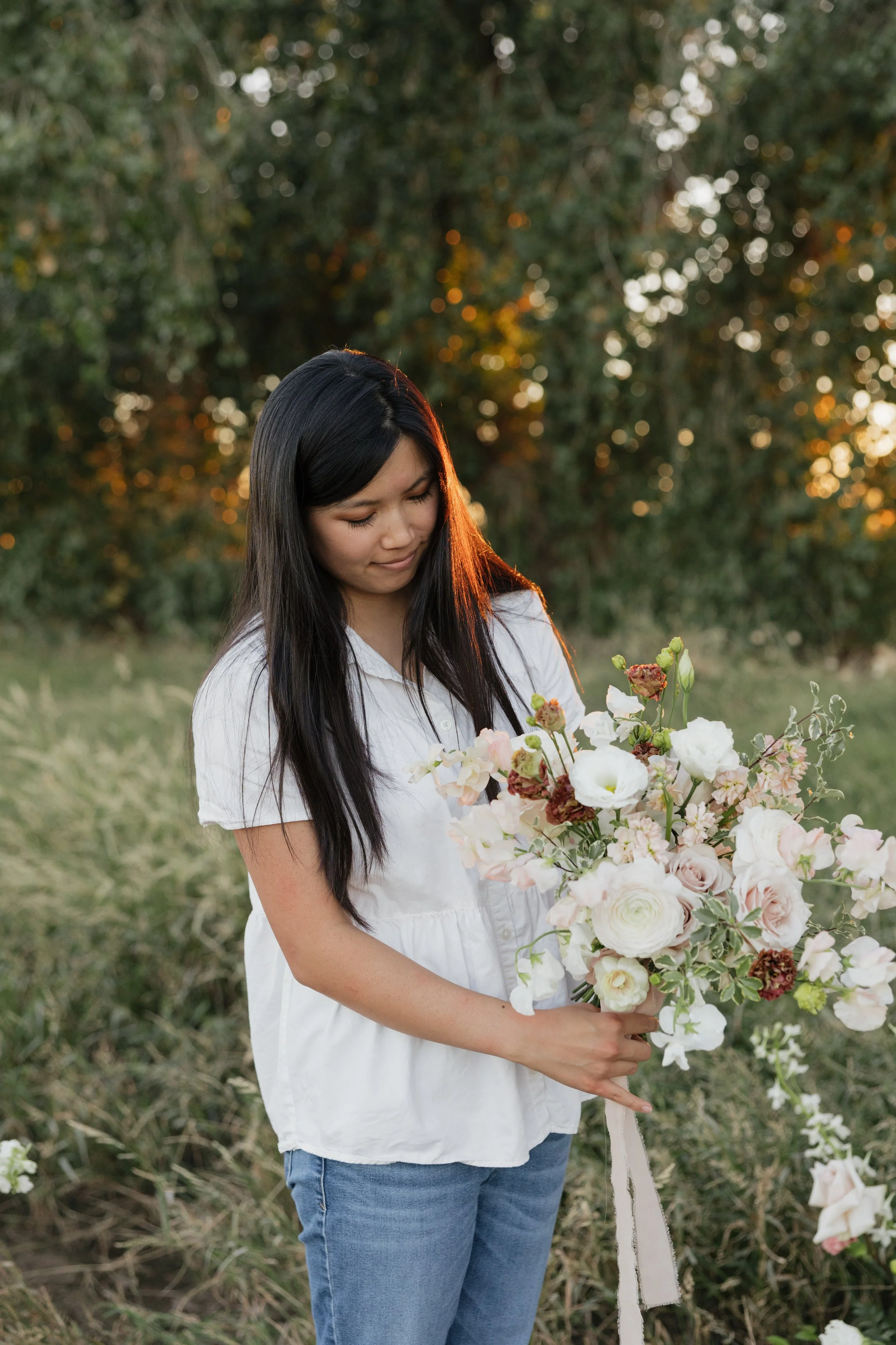A woman with long black hair and wearing a white blouse holding a bouquet of light pink and cream flowers outdoors during sunset.