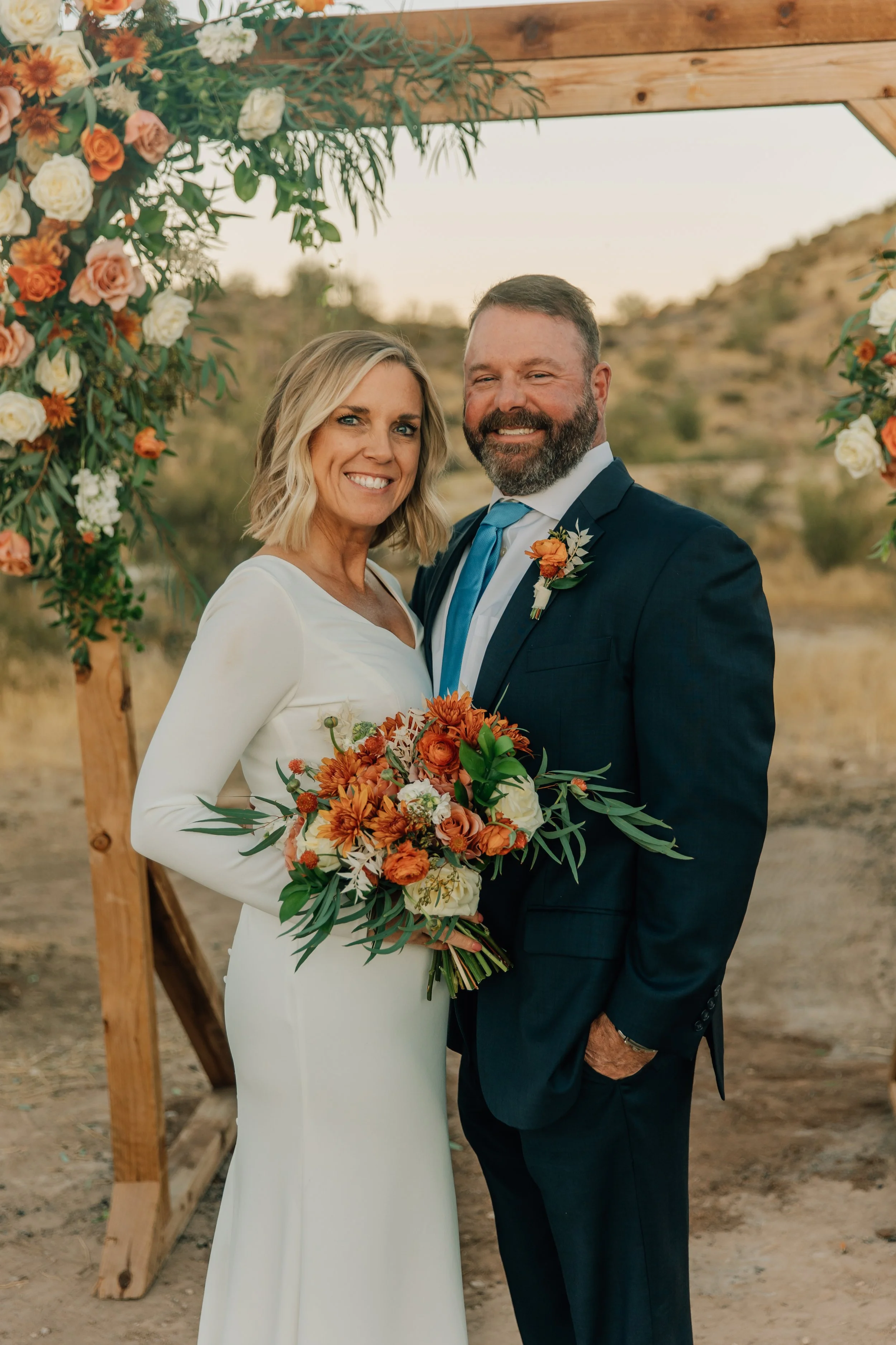 A bride and groom standing outdoors during sunset, smiling and holding a bouquet of orange, white, and green flowers, under a wooden arch decorated with similar floral arrangements.