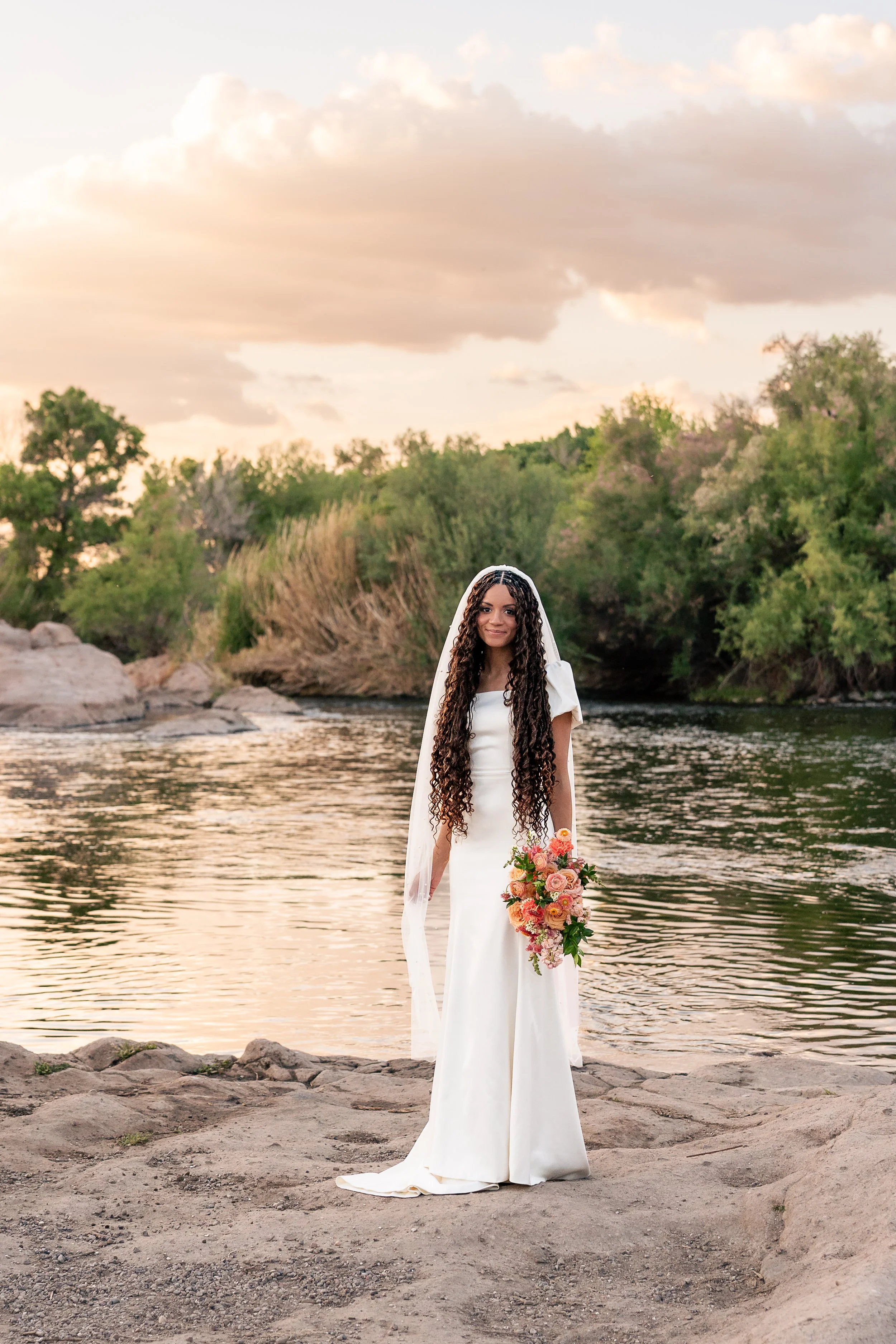 Bride standing near a river during sunset, holding a colorful bouquet, wearing a white wedding dress and veil.