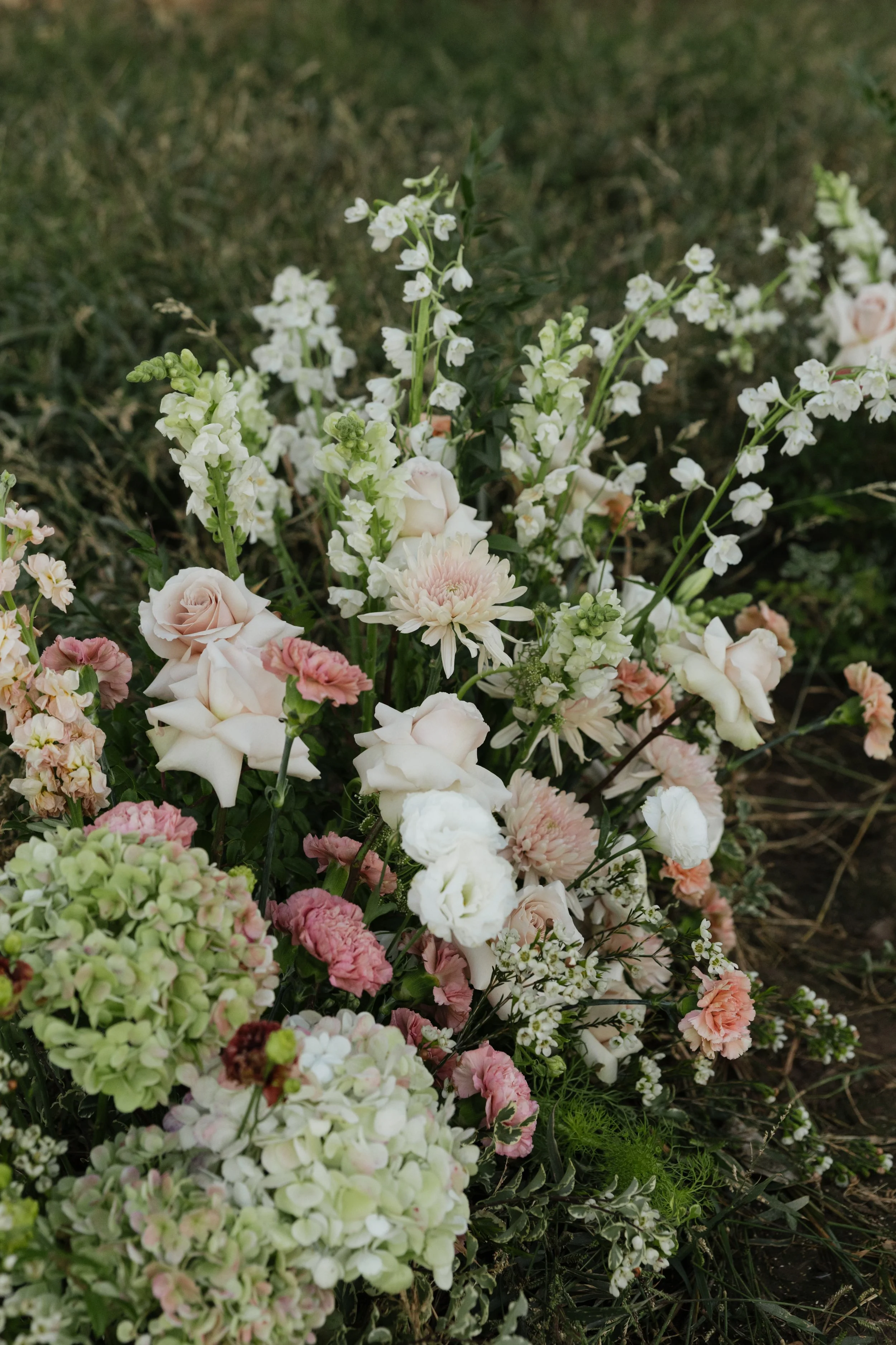 A bouquet of pale pink and white flowers, including roses, chrysanthemums, and other blossoms, arranged outdoors on grass.