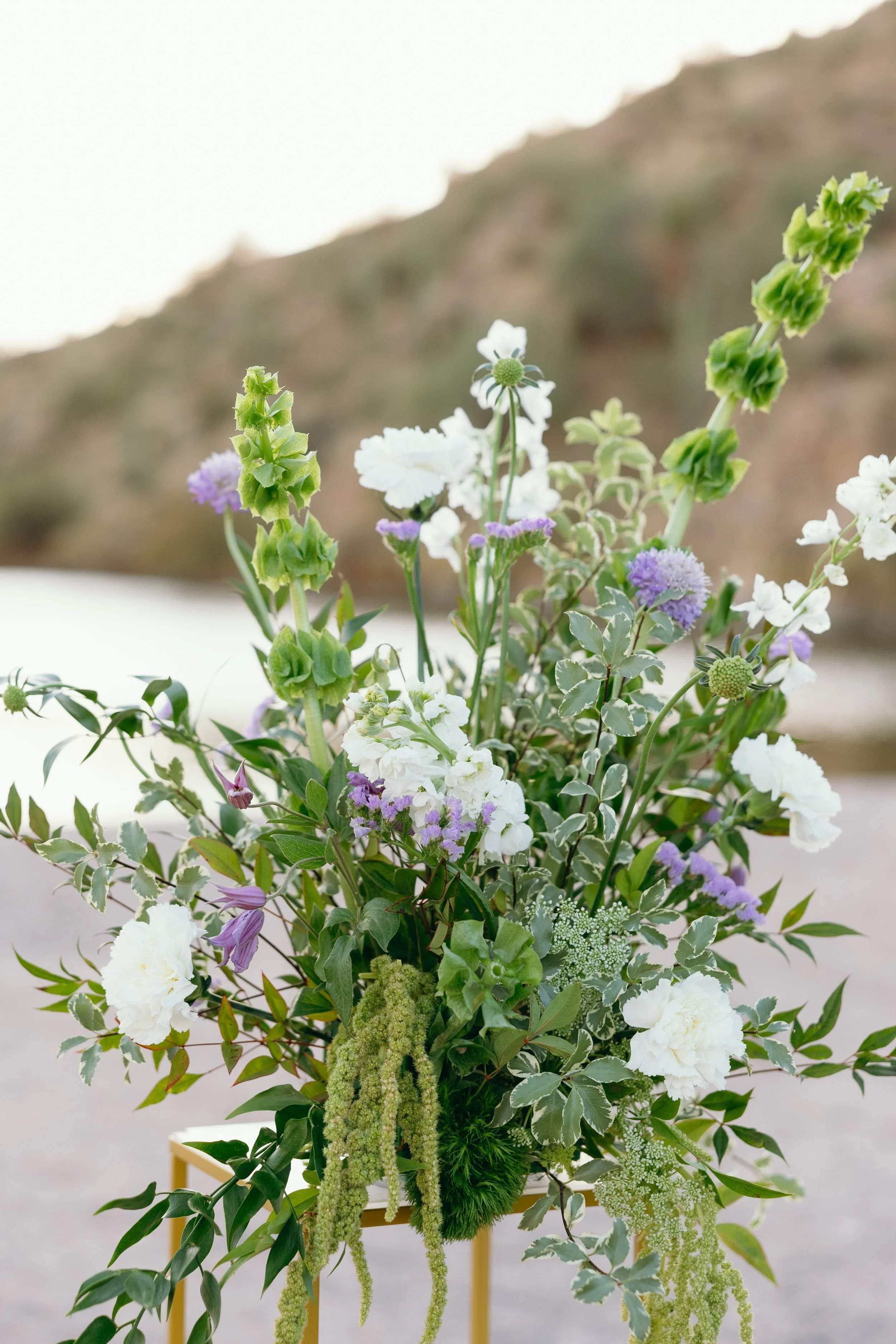 A standing arrangement of various flowers including white, purple, and green blooms on a yellow metal stand outdoors with a blurred landscape in the background.