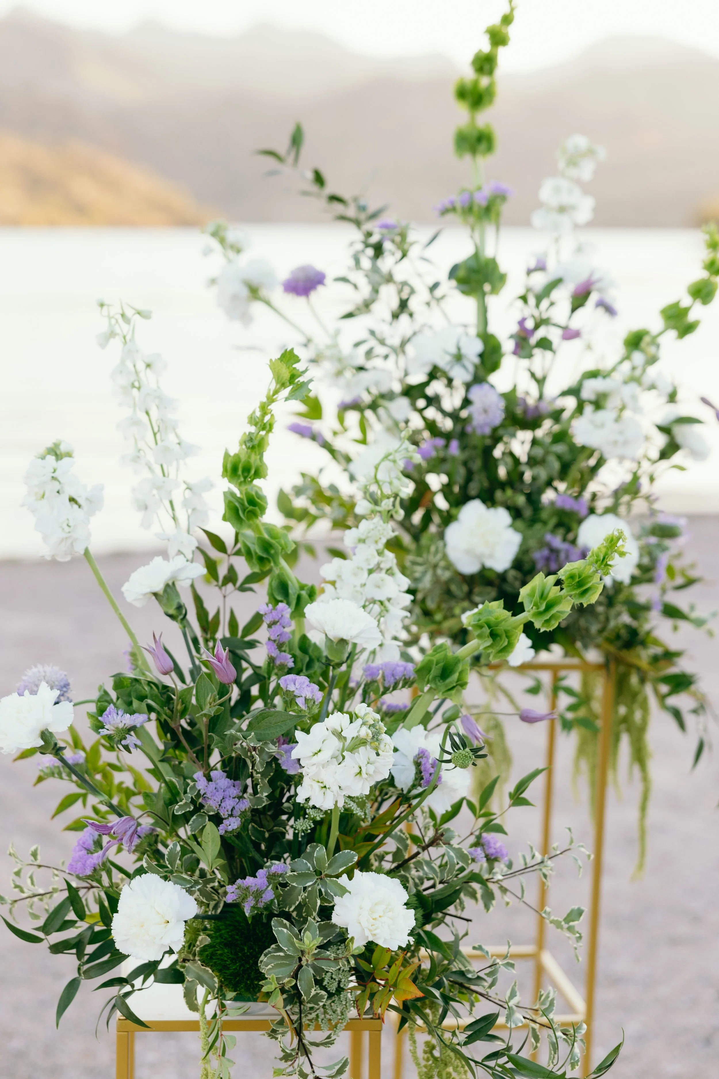 Two floral arrangements with white, purple, and green flowers on gold stands outdoors with mountains and a body of water in the background.